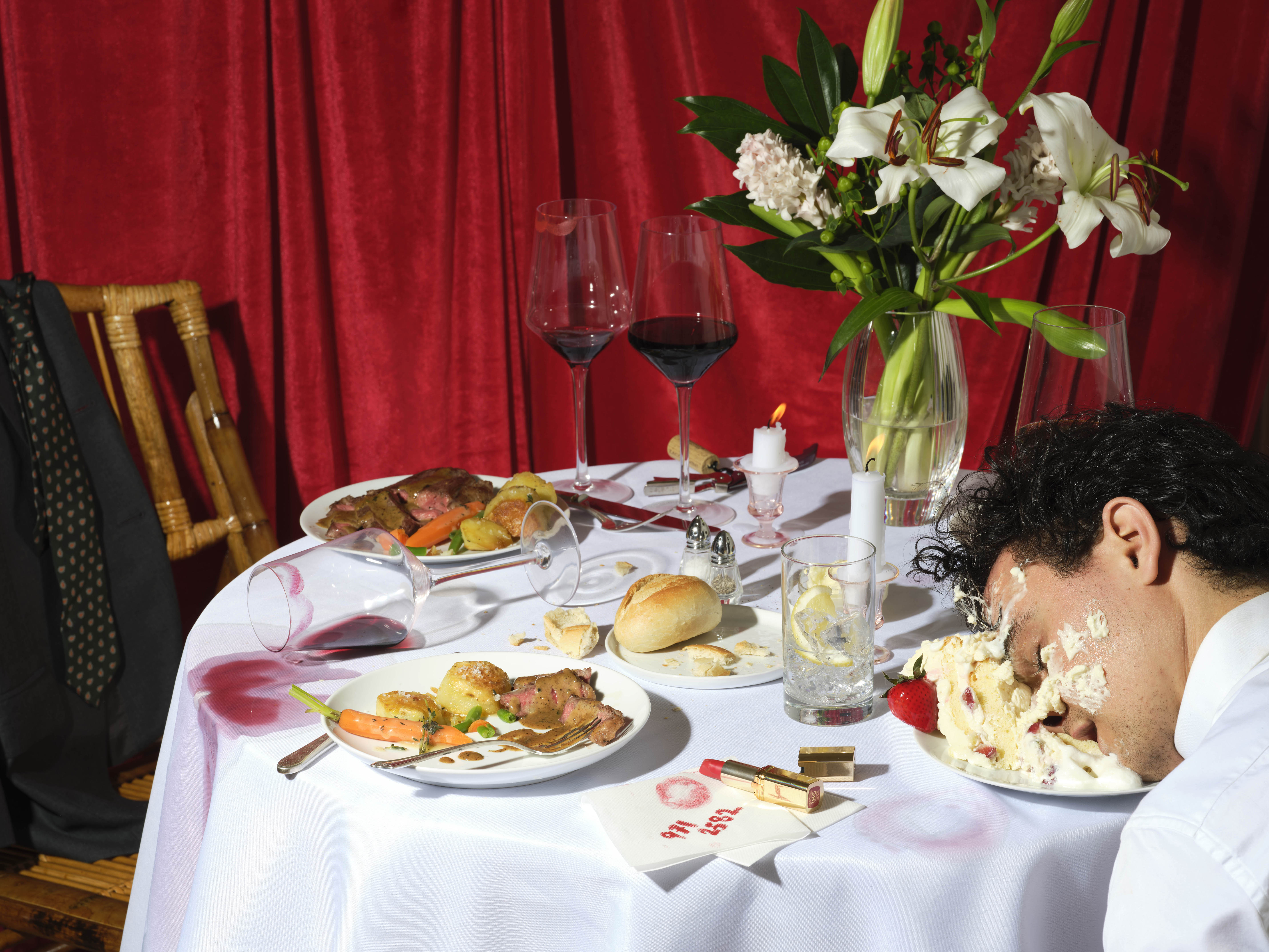 A table in the aftermath of a wedding party; there is spilled wine, half eaten plates of food, and a guess passed out in a slice of cake.