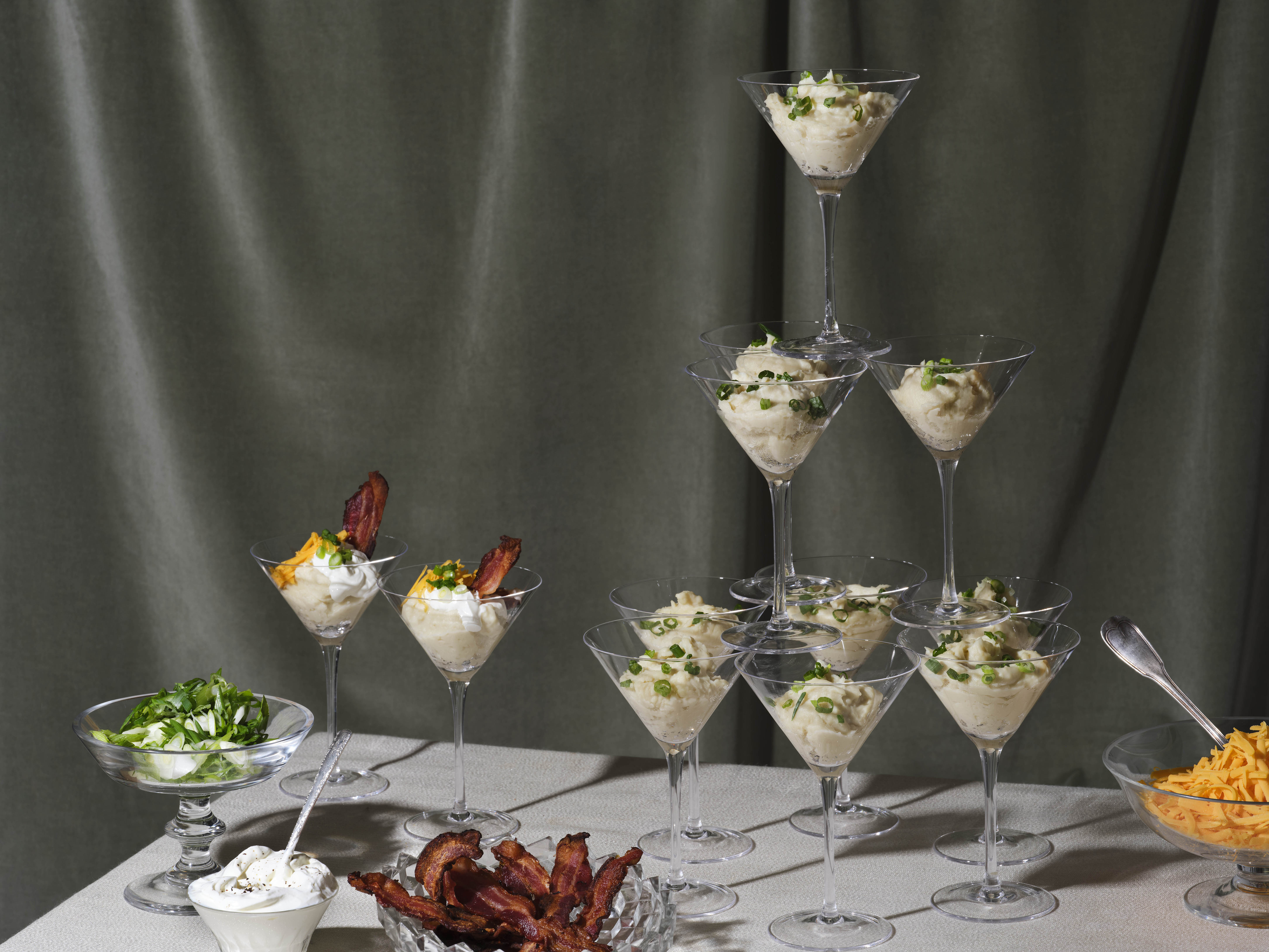 A tower of martini glasses filled with mashed potatoes topped with chives alongside a serving bowls filled with bacon, cheese, and sour cream.