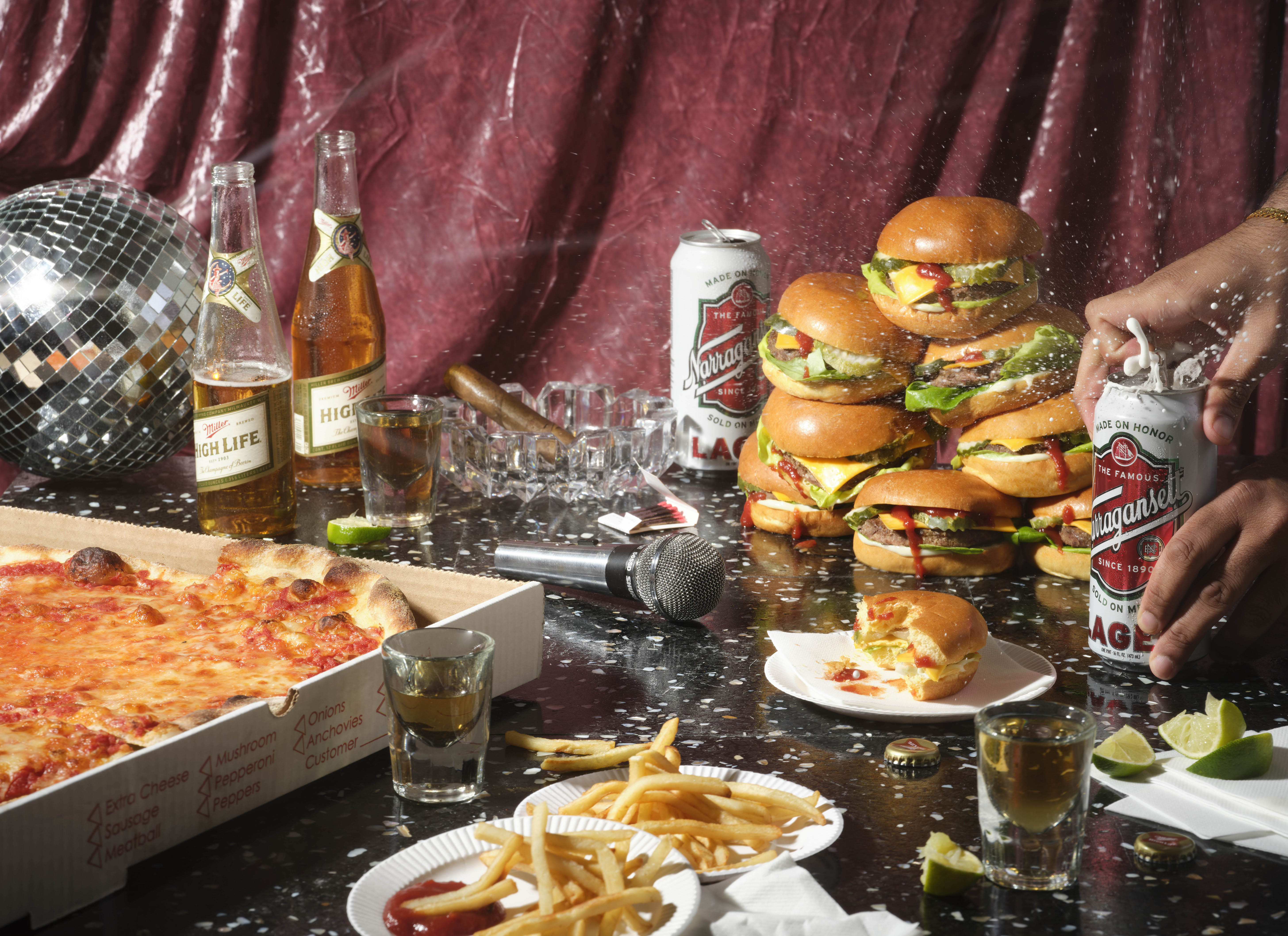A table mid-party, with a pizza, stack of burgers, french fries, disco ball, cigar, karaoke mic, and bottles and cans of beer.