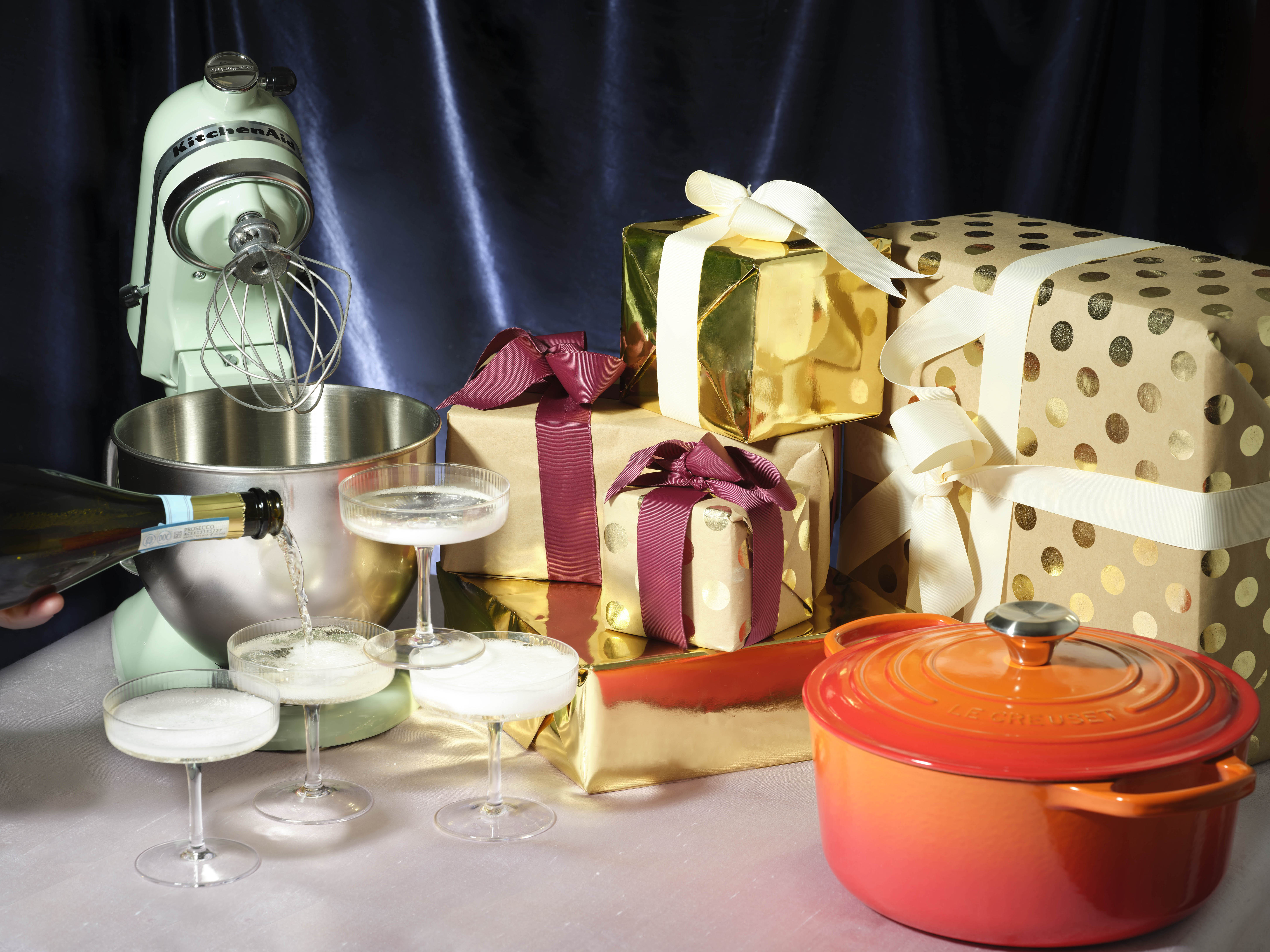 A table with wrapped gifts, along with a KitchenAid mixer, Le Creuset Dutch oven, and four full Champagne coupes.