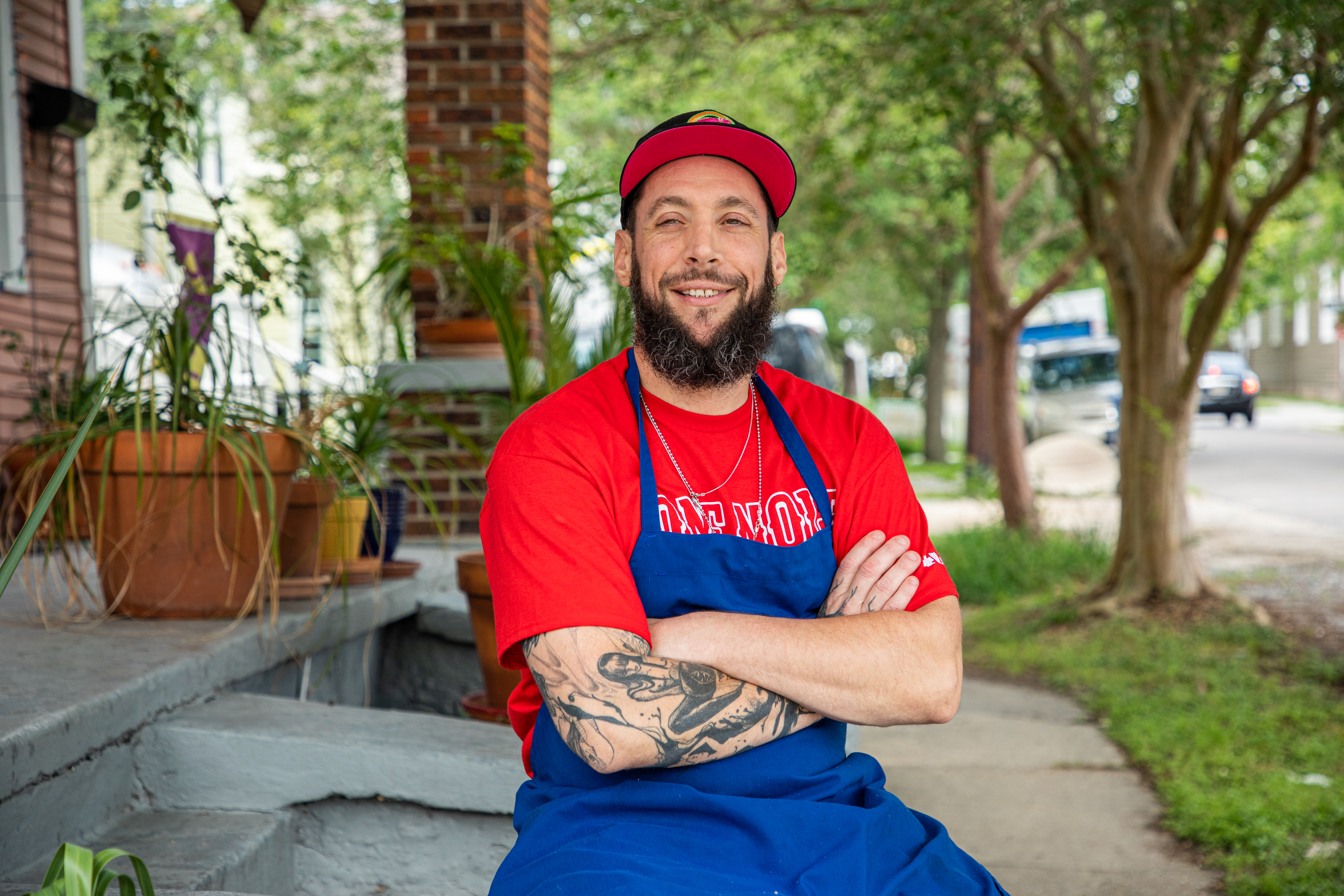 A smiling white man with one tattooed forearm sits with his arms crossed on a stoop wearing a red t-shirt and blue apron.