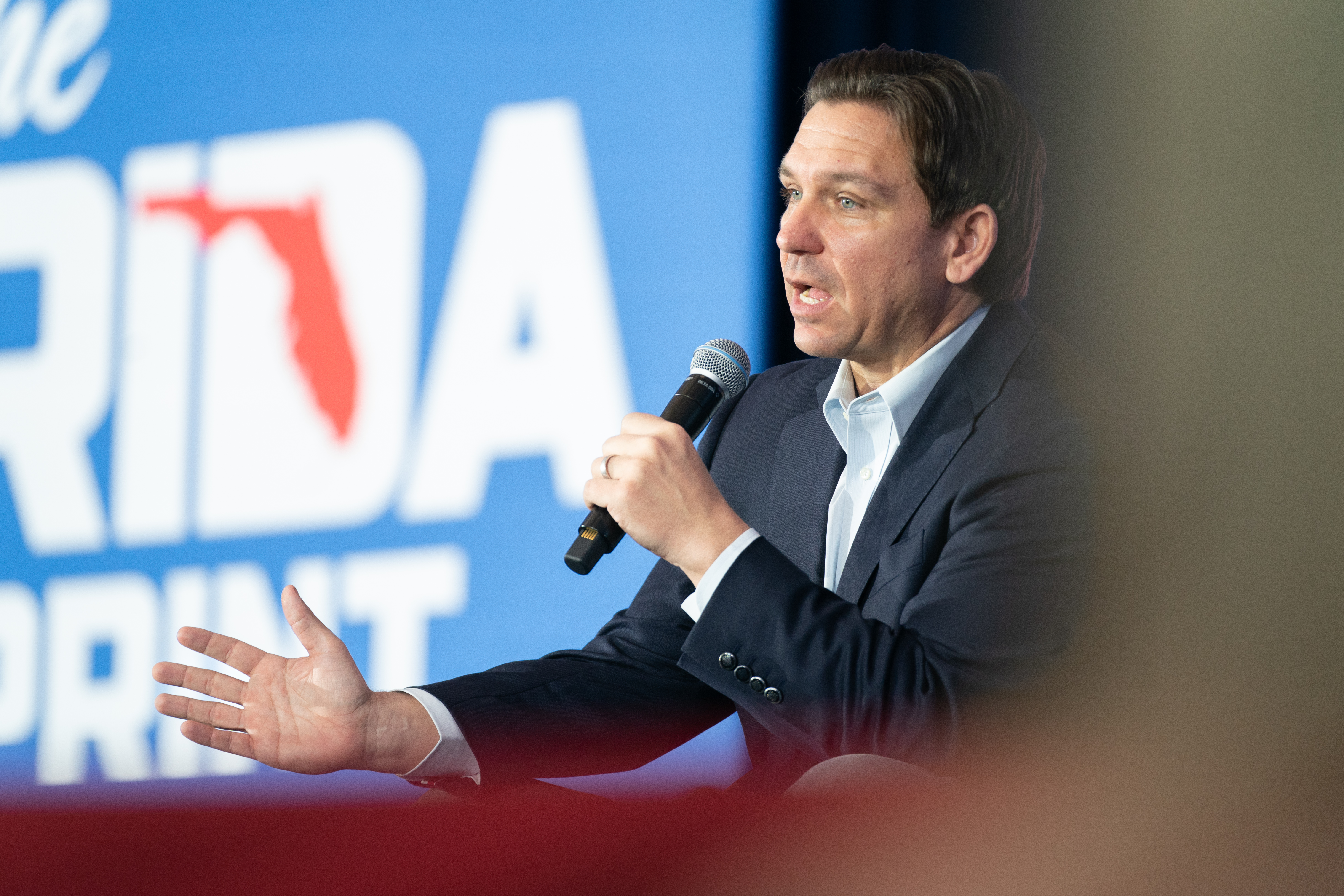 Ron DeSantis speaks into a handheld microphone, gesturing with his other hand. He’s wearing a blue suit and white shirt, and stands in front of a tall blue backdrop showing the state of Florida.