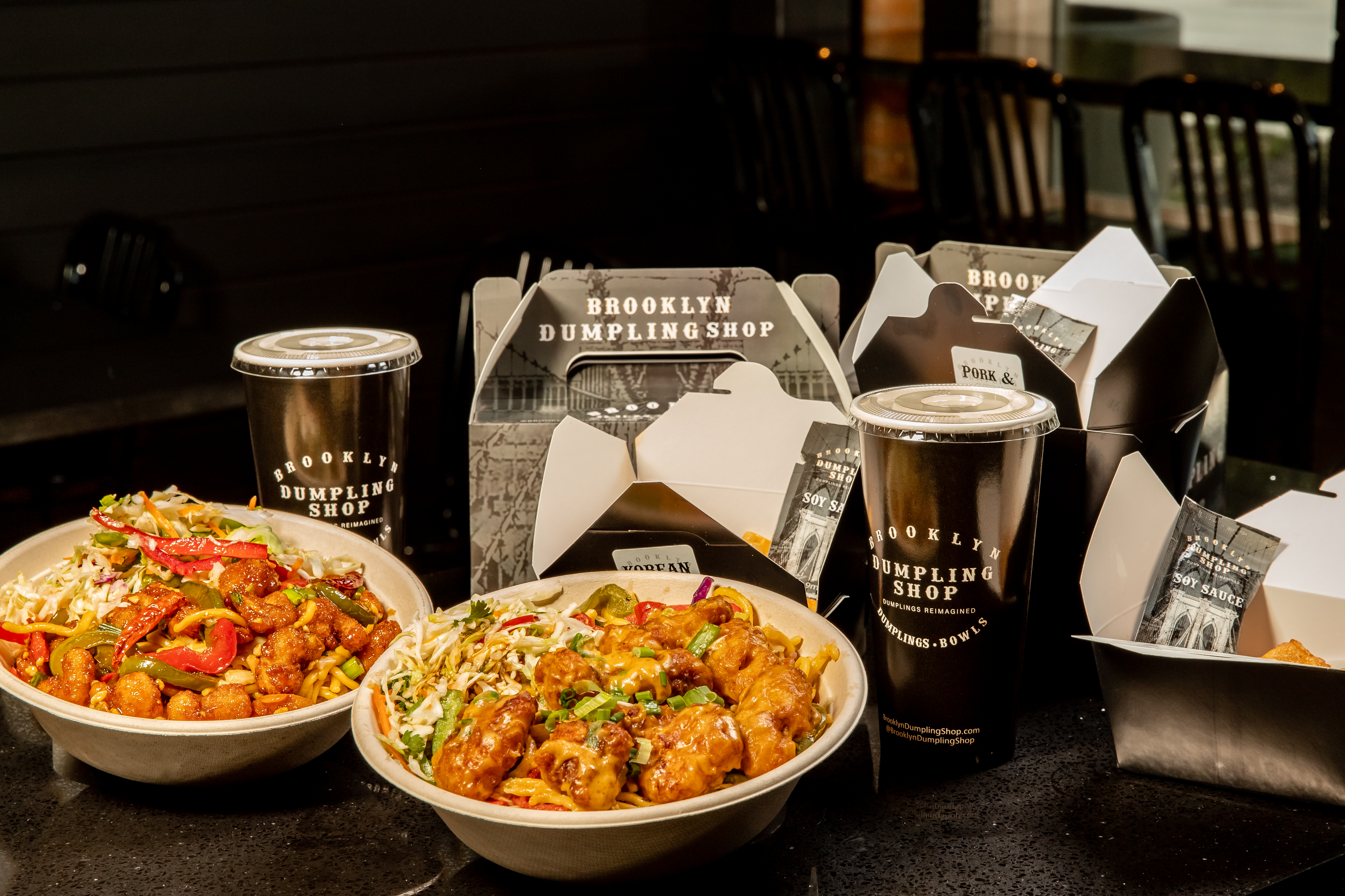 On a black background, bowls with assorted dumplings sit in front of black boxes embossed with a white logo reading “Brooklyn Dumpling Shop.”
