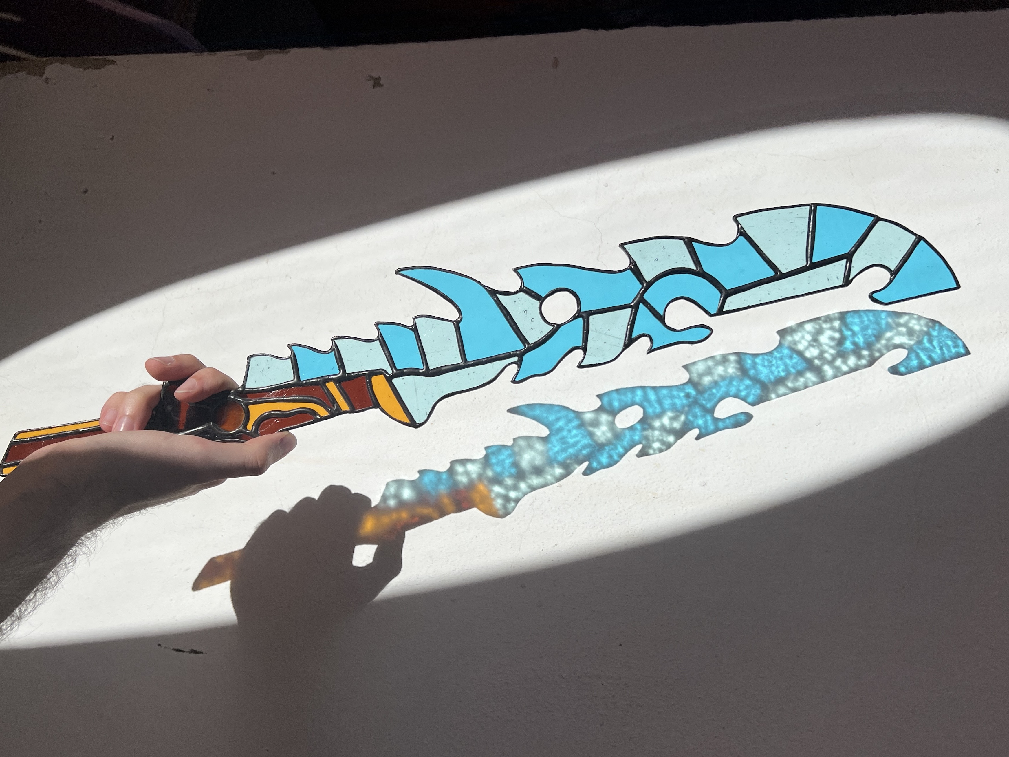 A Guardian Sword++ from Zelda: Breath of the Wild, but made out of stained glass. It’s being held in a spotlight over a white surface, so its colorful shadow can be seen below it.