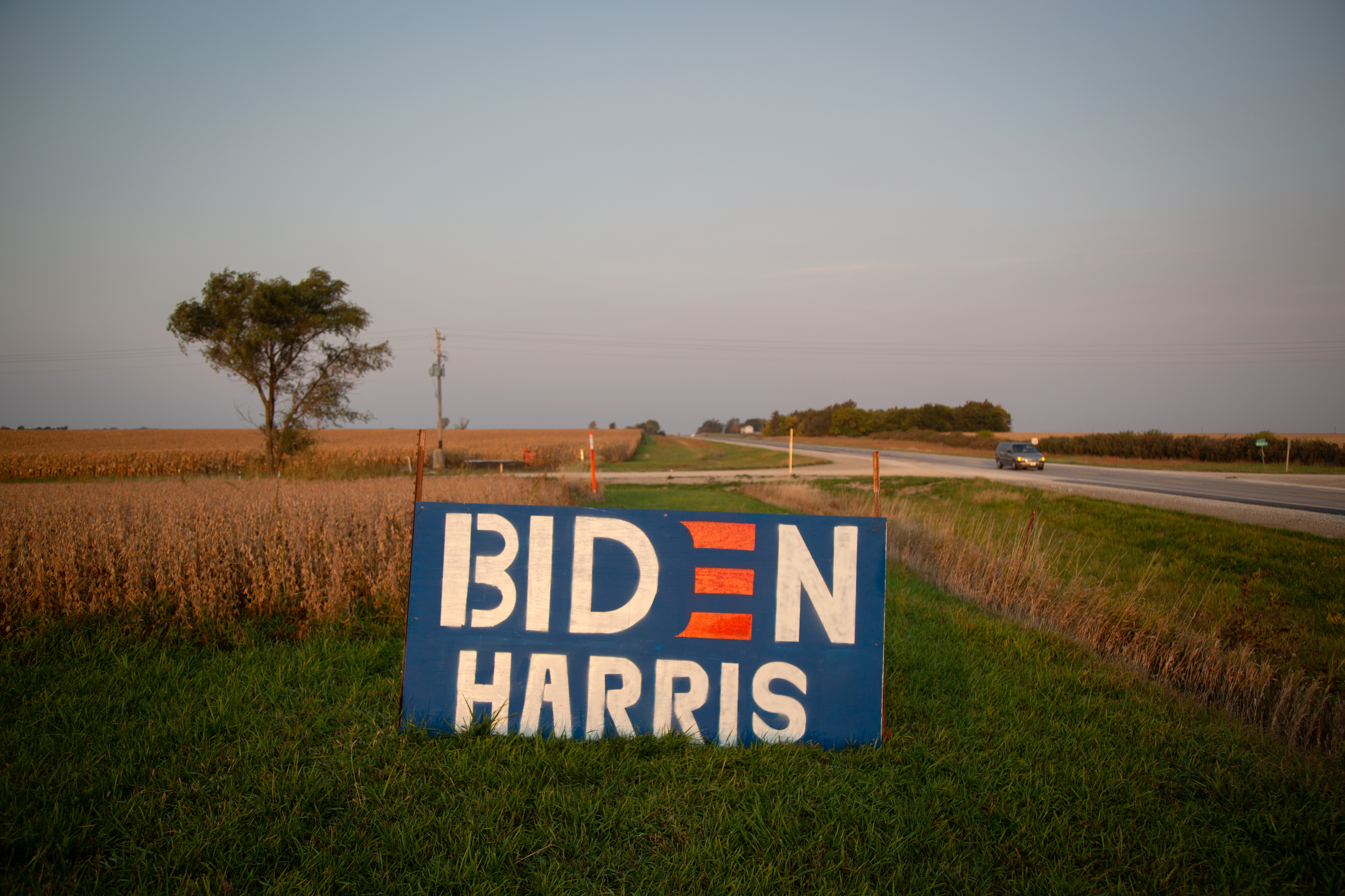 A large red and blue Biden-Harris sign sits on the side of a dirt road in Iowa, with farm fields stretching out behind it.