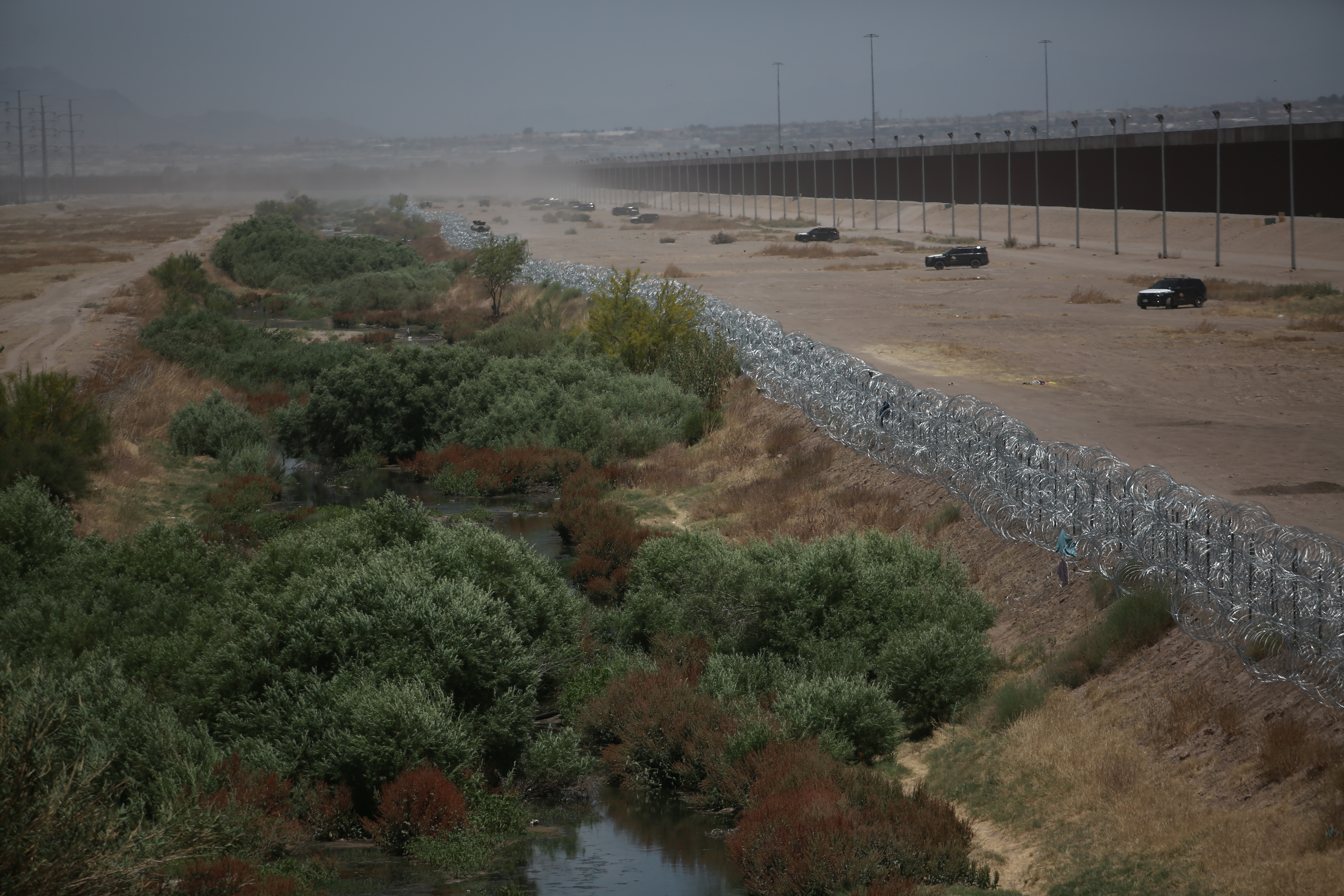 US-Mexico border comprised of green, desert shrubs and pond water, a metal fence, and sand. SUVs patrol the border, parked in front of a tall cement wall lined with floodlights