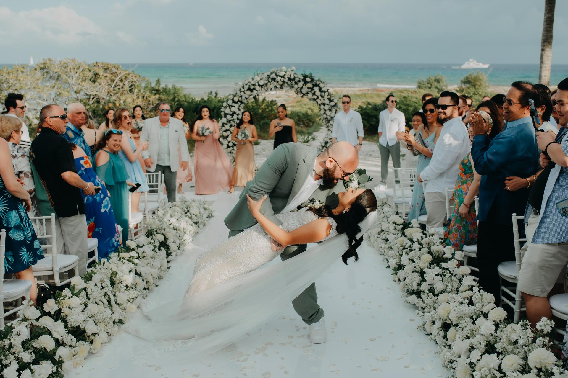 A beaming groom embraces his bride and leans her back for a kiss after saying, “I do,” while a clapping crowd watches. A sandy beach and white floral structures shine in the afternoon sun behind them.
