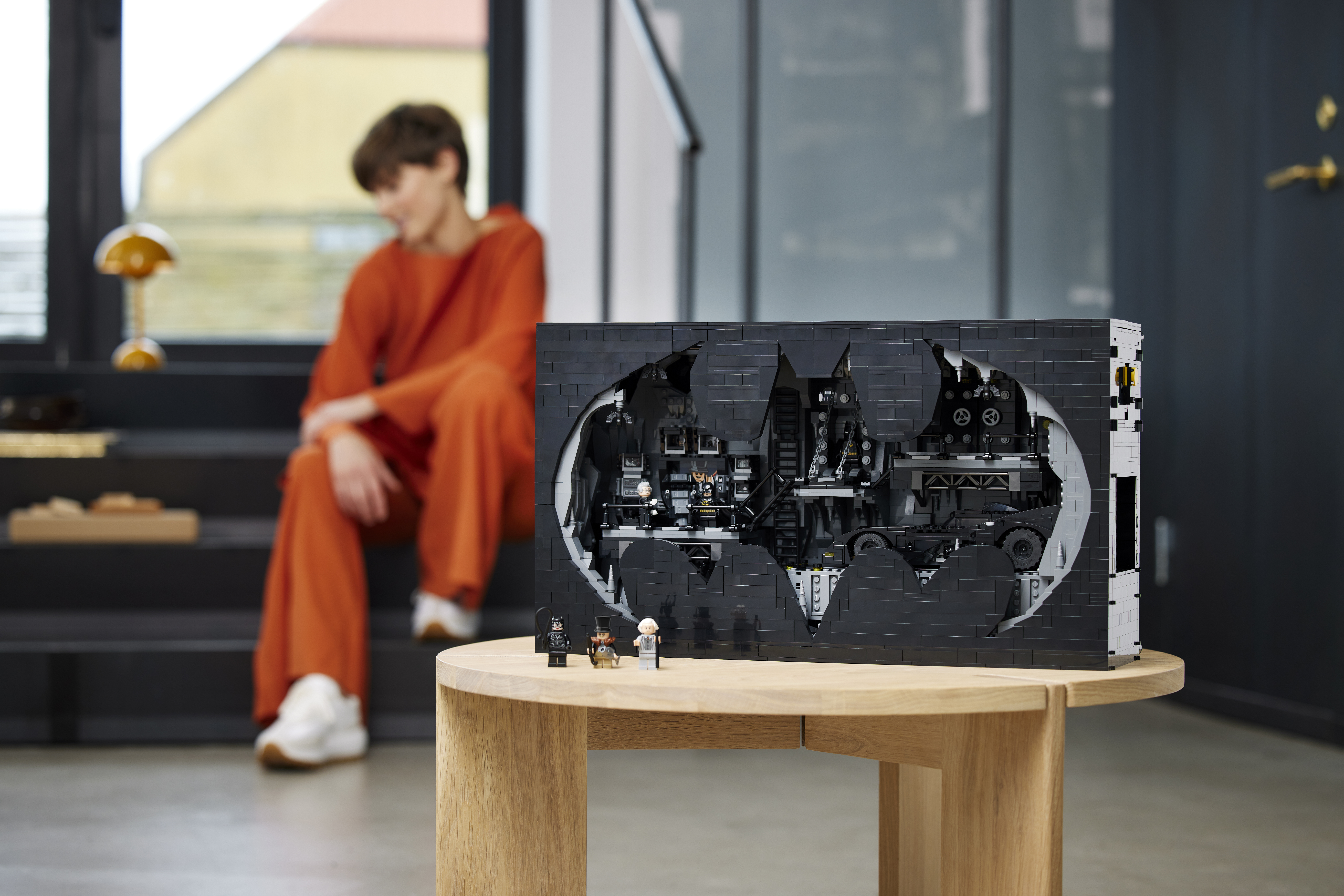 Lego’s Batcave Shadow Box set sits closed on a wooden table. It’s shaped like a wide, tall box, with a “cutout” of the batsymbol on the front, through which can be seen an intricate Lego diorama of the Batcave from Batman 1989. A person wearing orange sits in the background. 