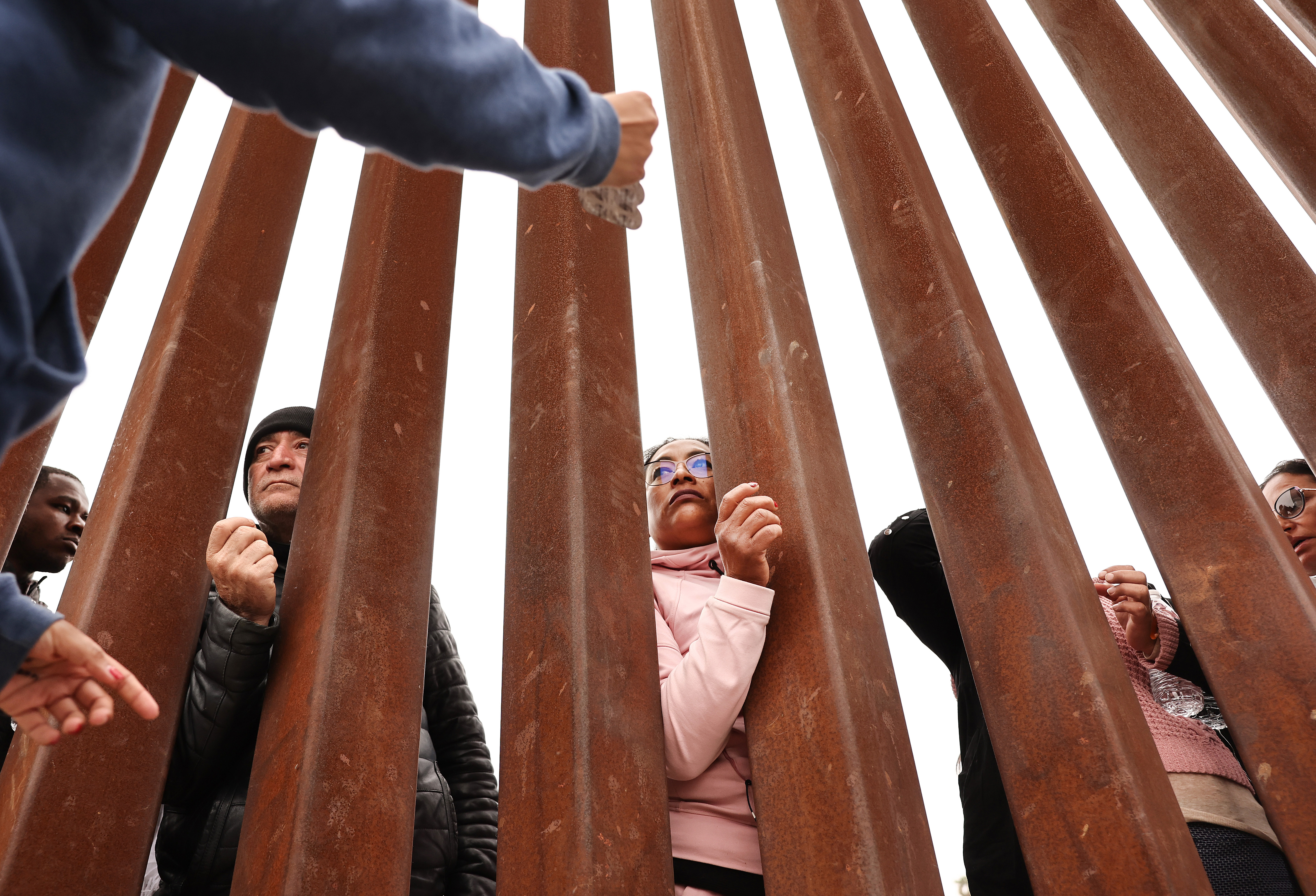 Several people look through the upright metal columns of a border wall as volunteers talk to them from the other side.