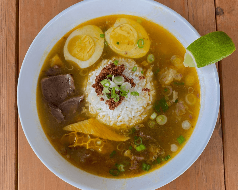 A bowl of broth, rice, sliced meats, halved boiled eggs, and a slice of lime on the rim.
