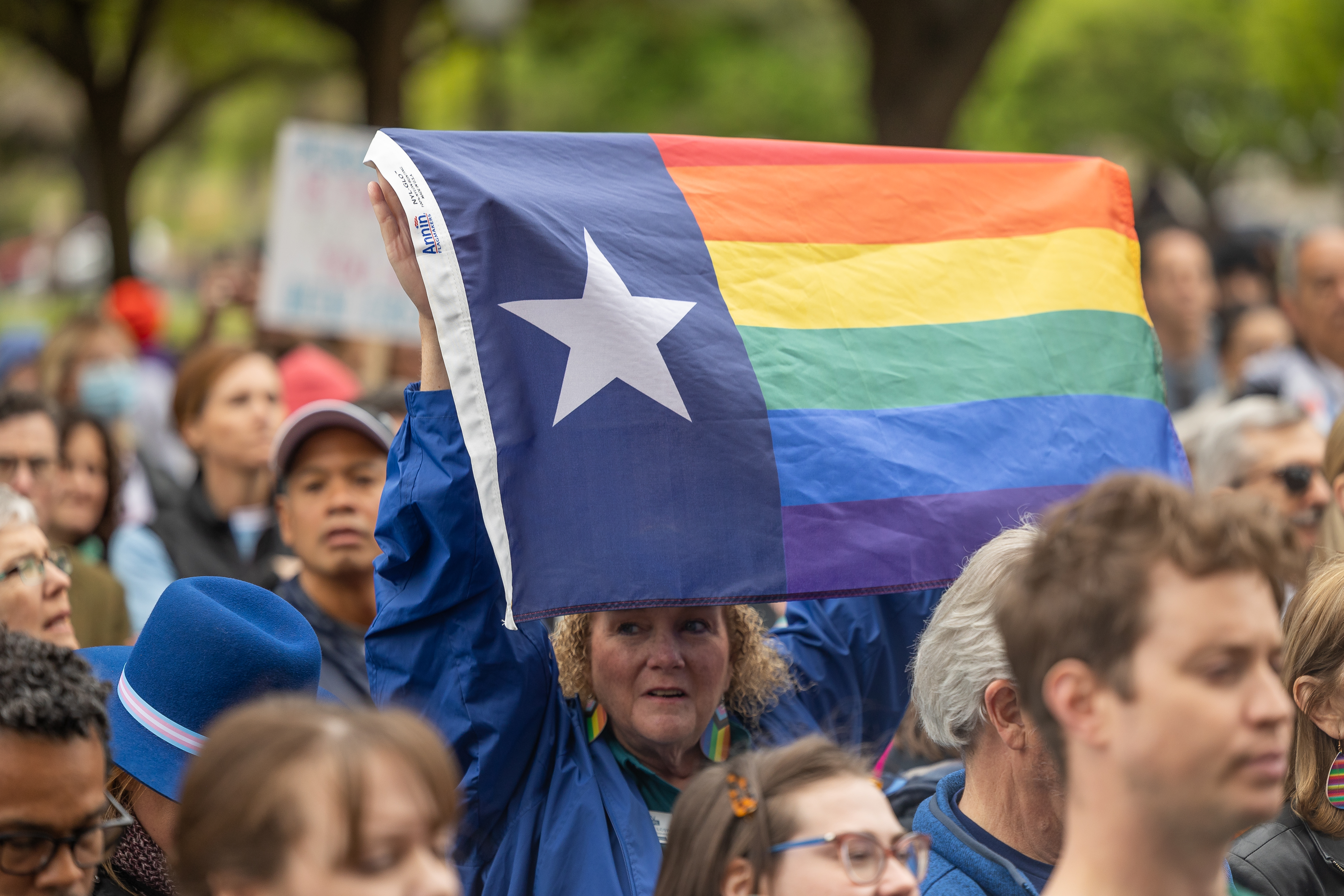 A crowd of people, some holding signs, with one in the center holding up a rainbow Texas flag.