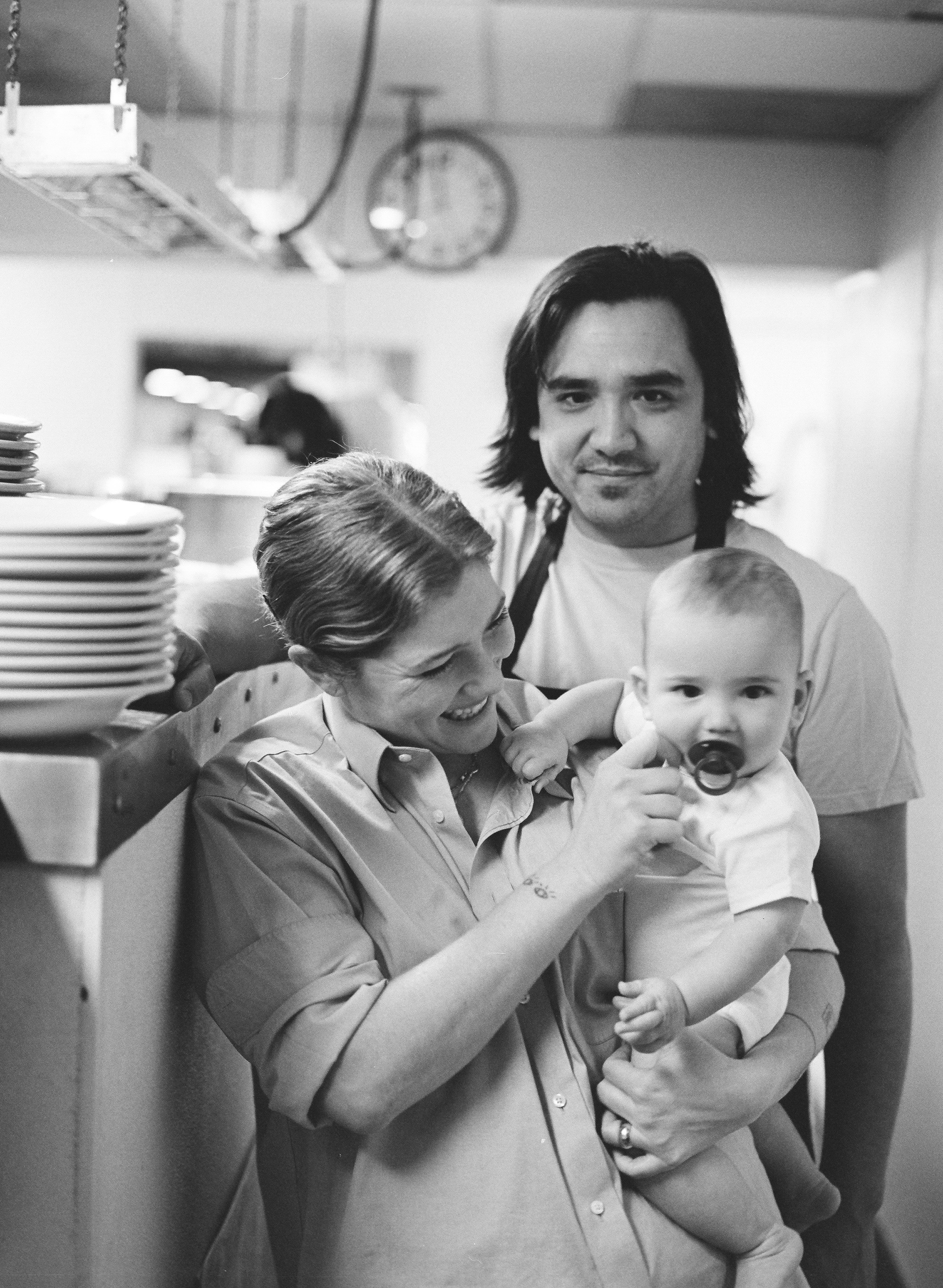 A woman with blonde curly hair holding a baby and a man with shoulder-length dark hair wearing an apron stand in a restaurant kitchen. 