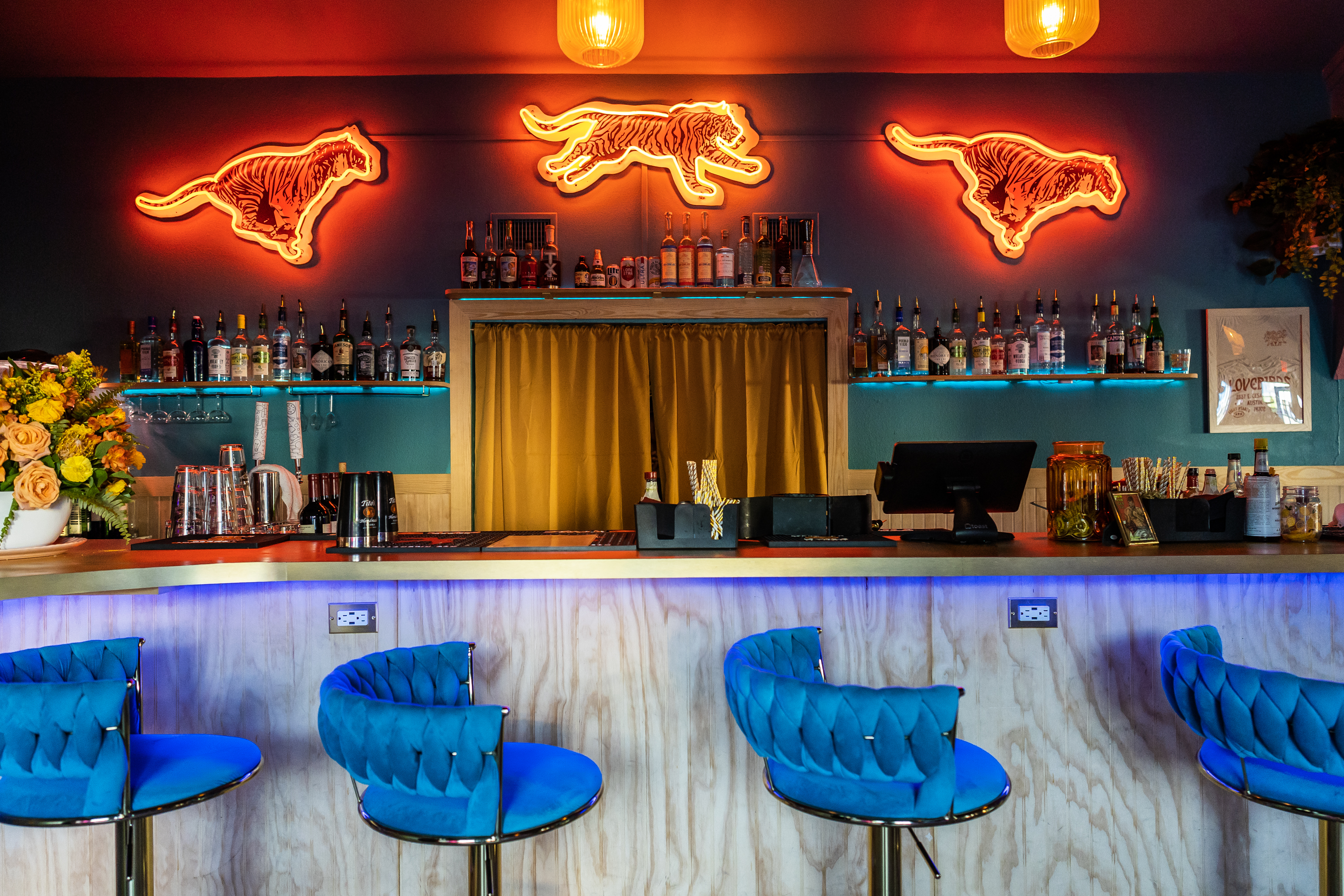 A cocktail bar with blue-clothed bar stools and neon tigers on the wall above the bottle shelves.