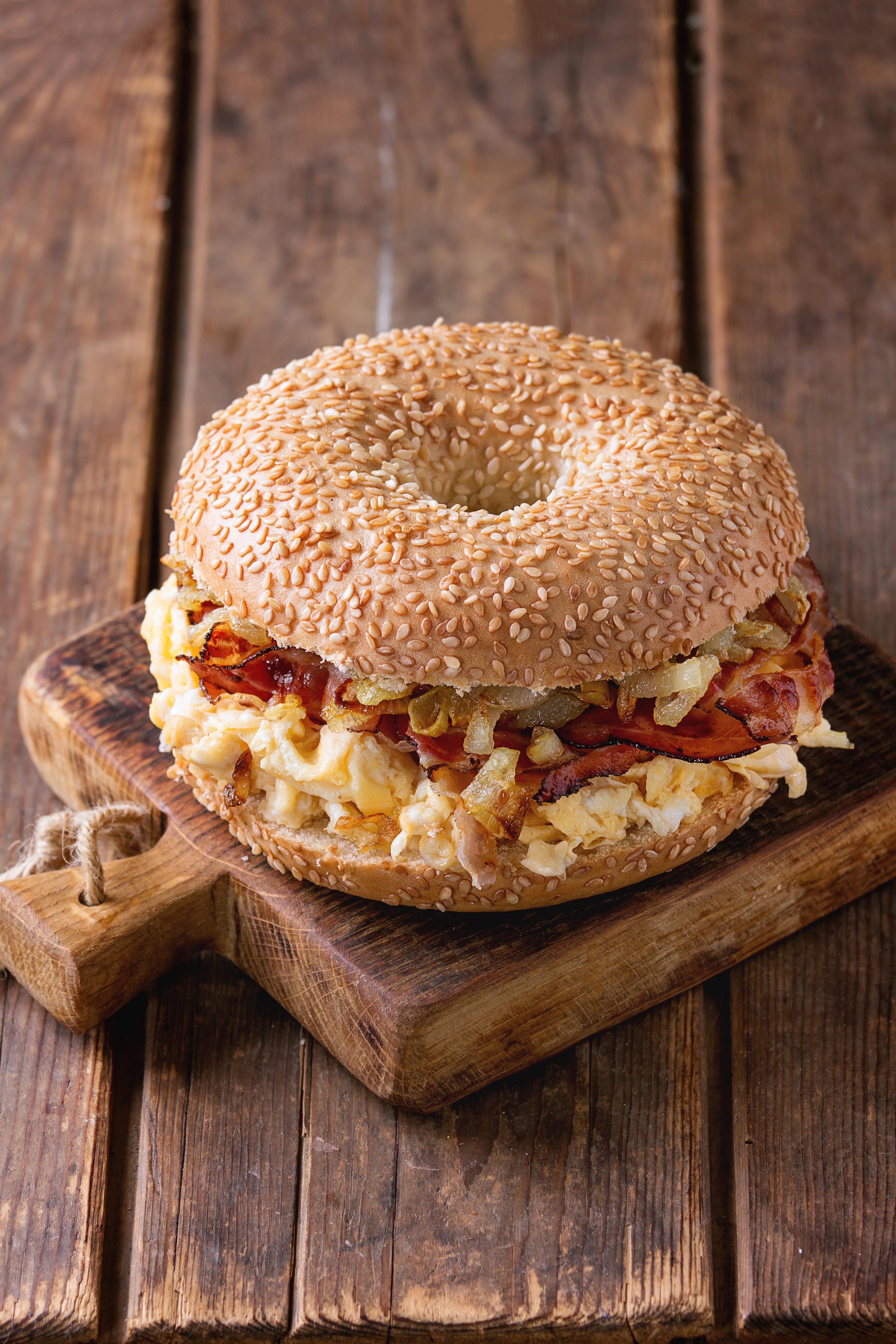 Bagel with bacon, scrambled eggs and fried onion on small wooden chopping board over wooden background.