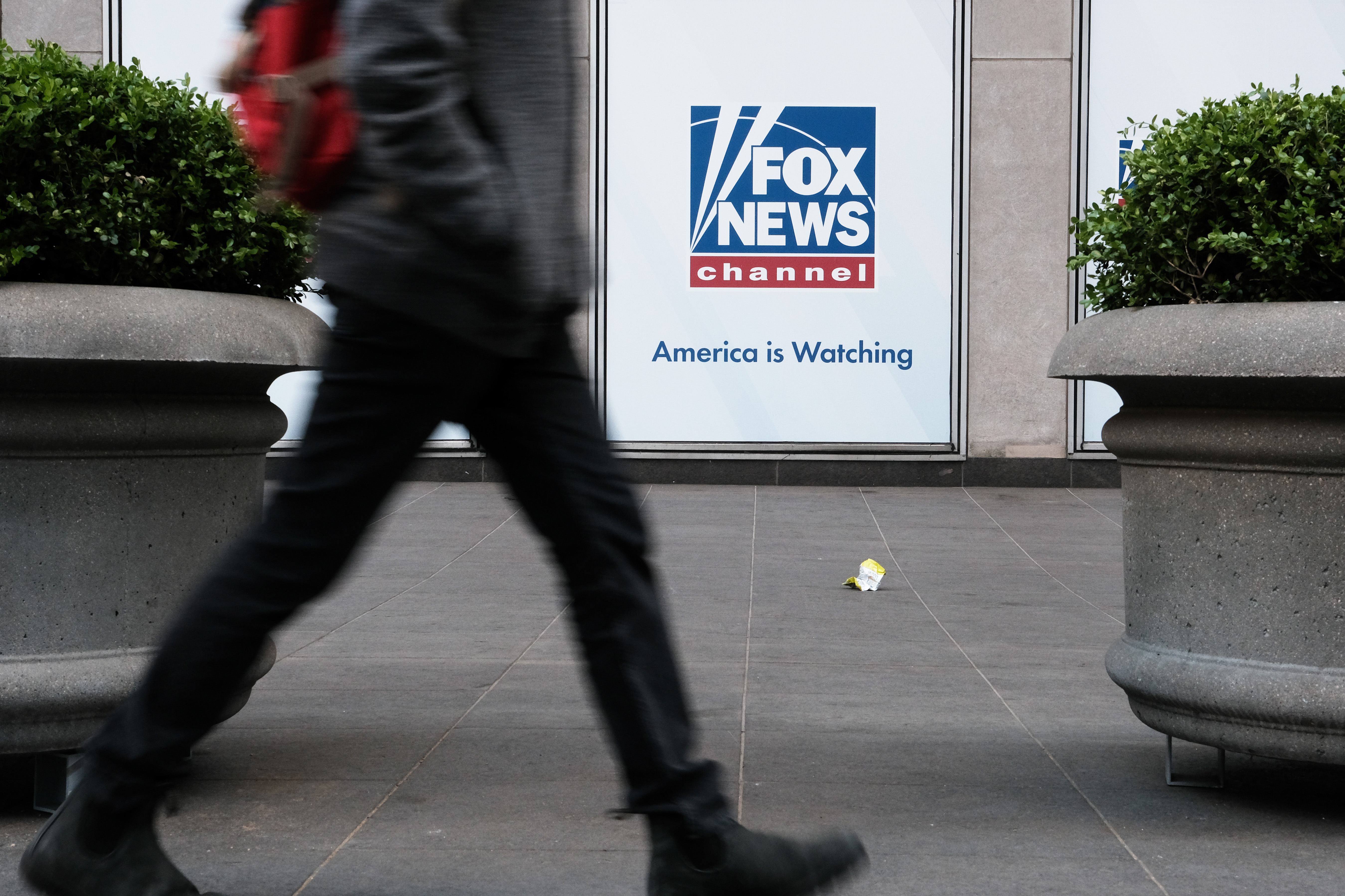 A person walks by the planters outside the News Corp headquarters in New York City, which is displaying a sign that reads “Fox News channel. America is watching.”