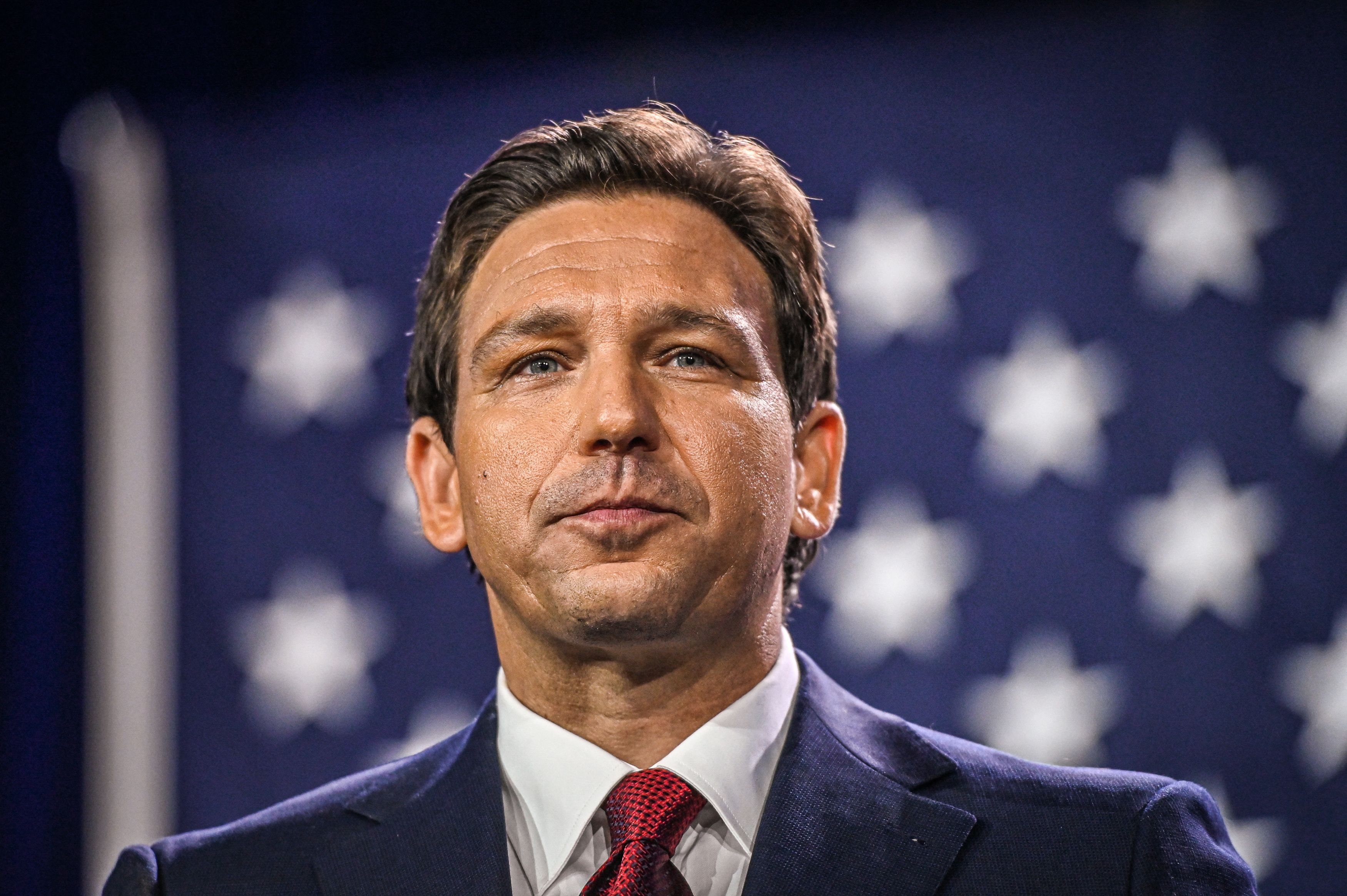 A portrait of DeSantis, clean shaven in a navy suit, red tie, and white shirt. He bears a serious expression, the US flag stretched out behind him.
