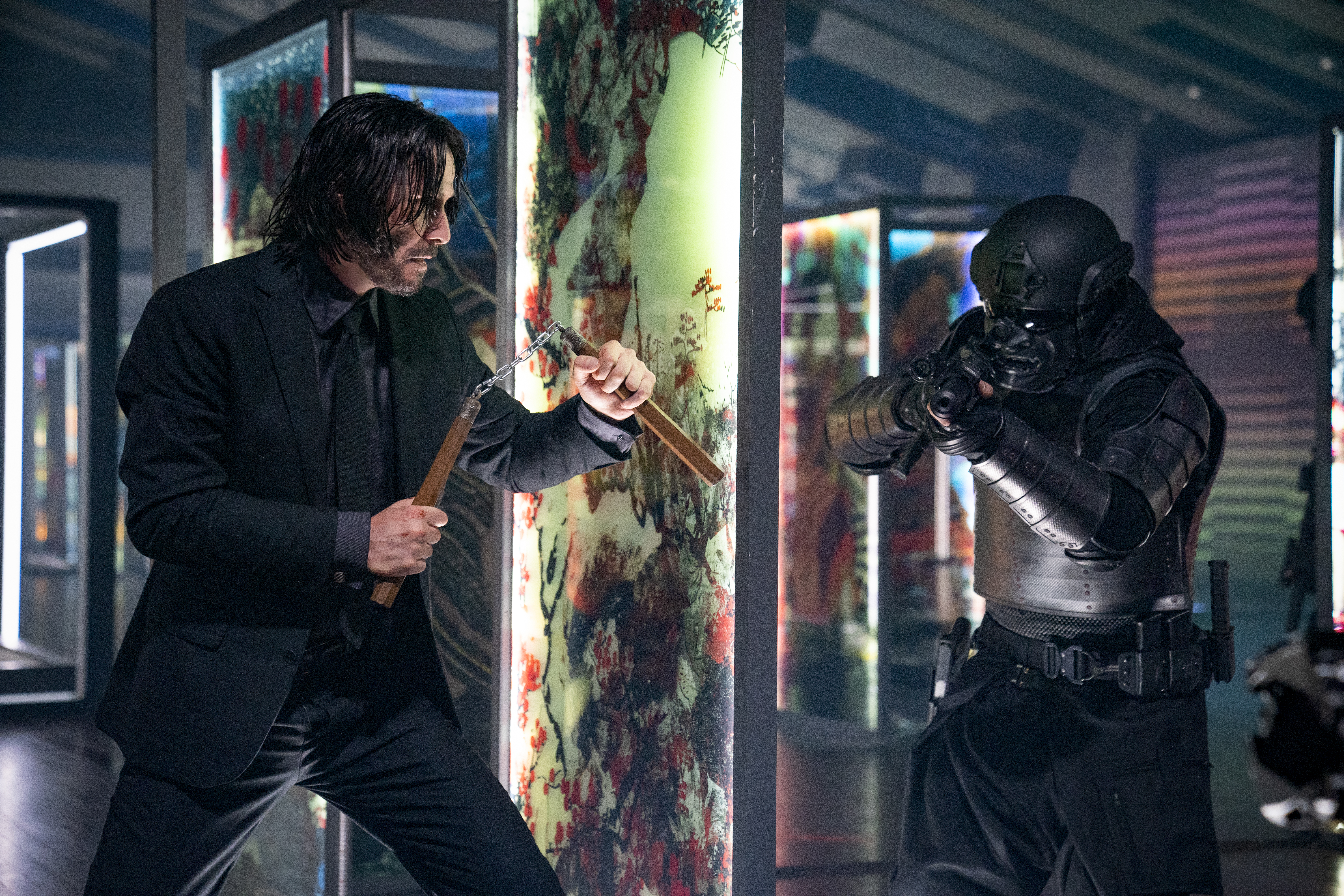 Keanu Reeves as John Wick holding num-chuks behind a glass panel preparing to ambush a soldier in black samurai armor in John Wick: Chapter 4.