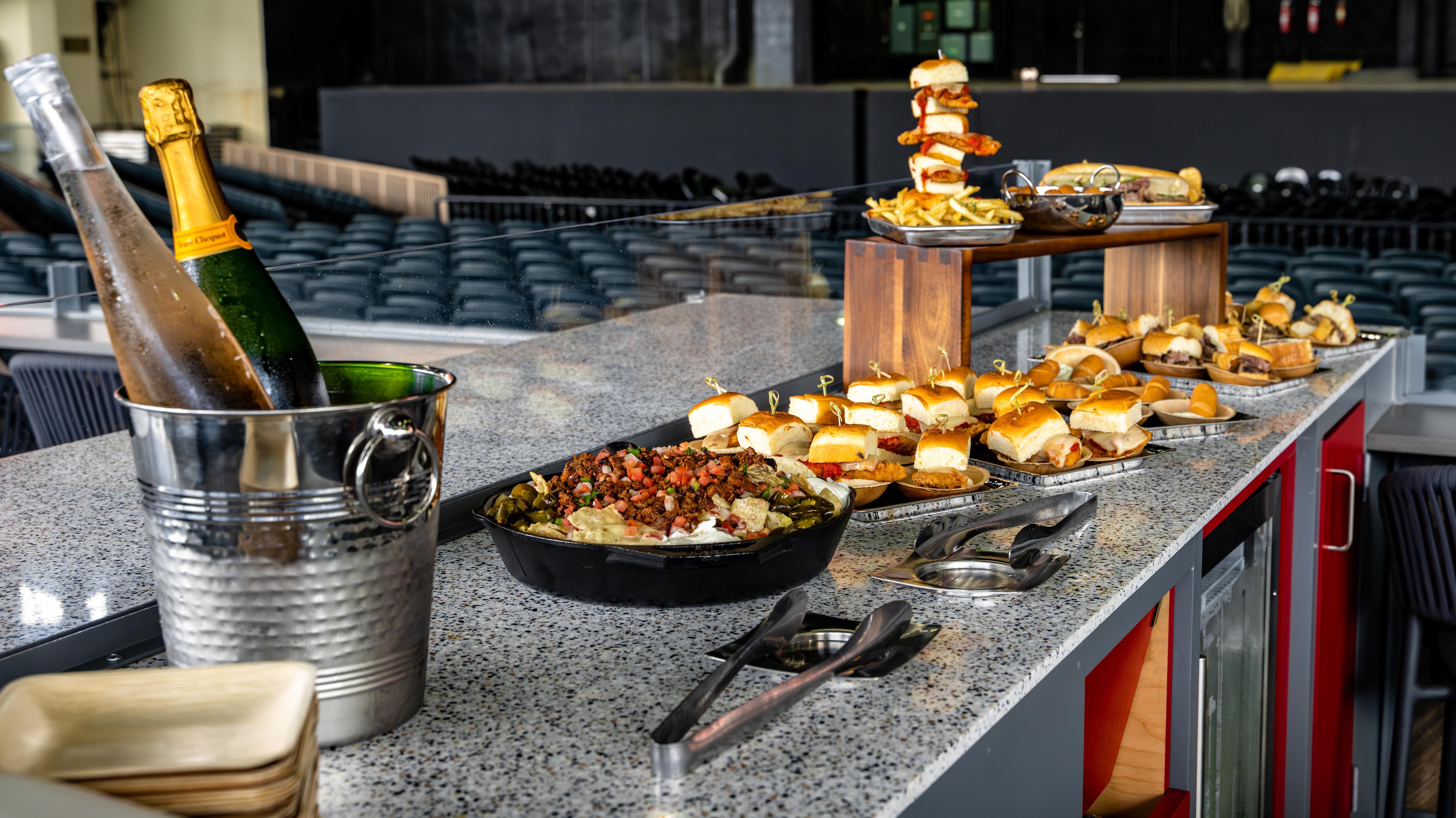 A table holds a bucket with two bottles of wine, nachos in a cast iron skillet, plates of sliders. In the background, there’s a stage in an amphitheater.