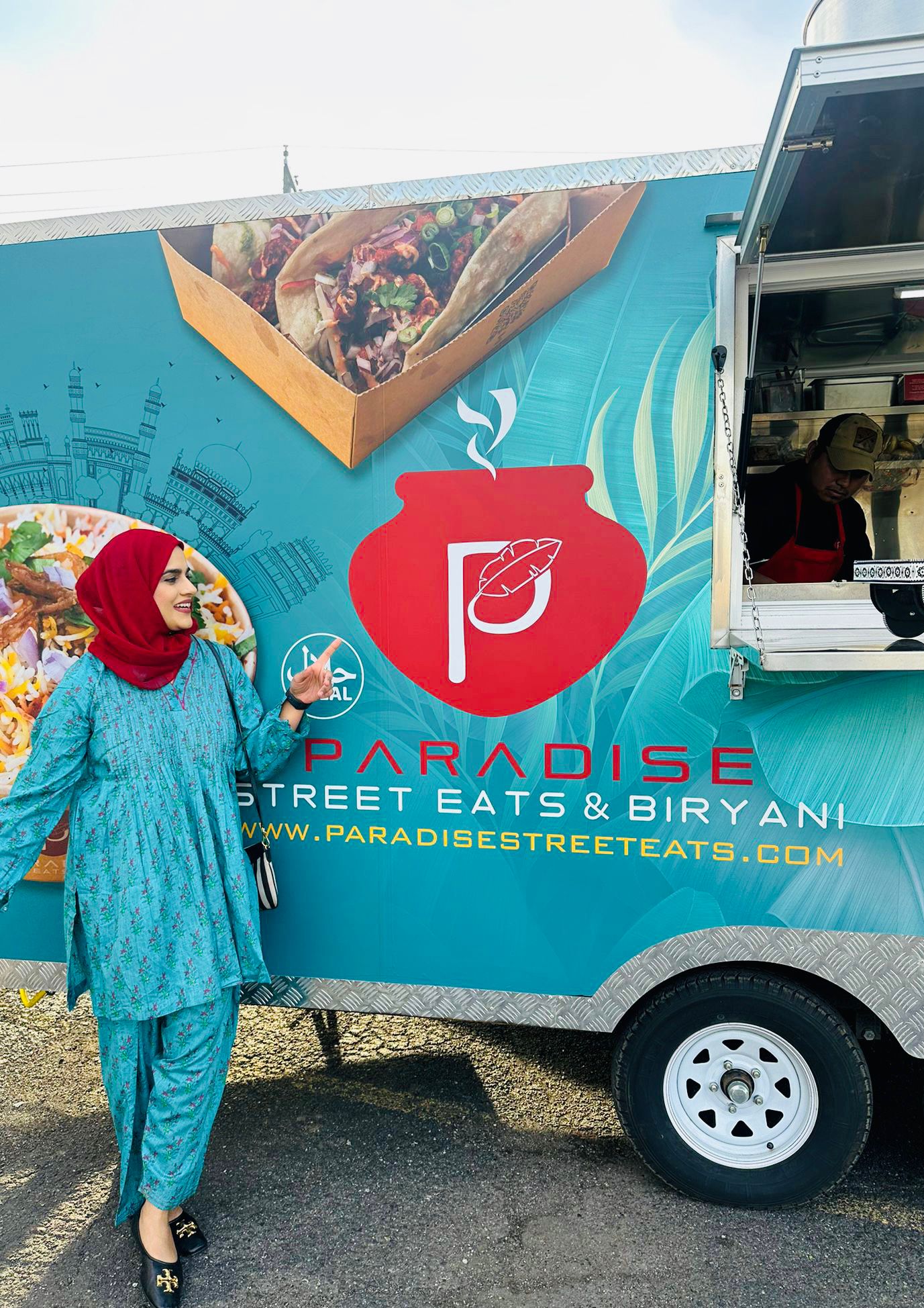 A woman in aqua blue dress and scarlet headscarf standing in front of a food truck in aqua blue and red that says Paradise Street Eats &amp; Biryani.