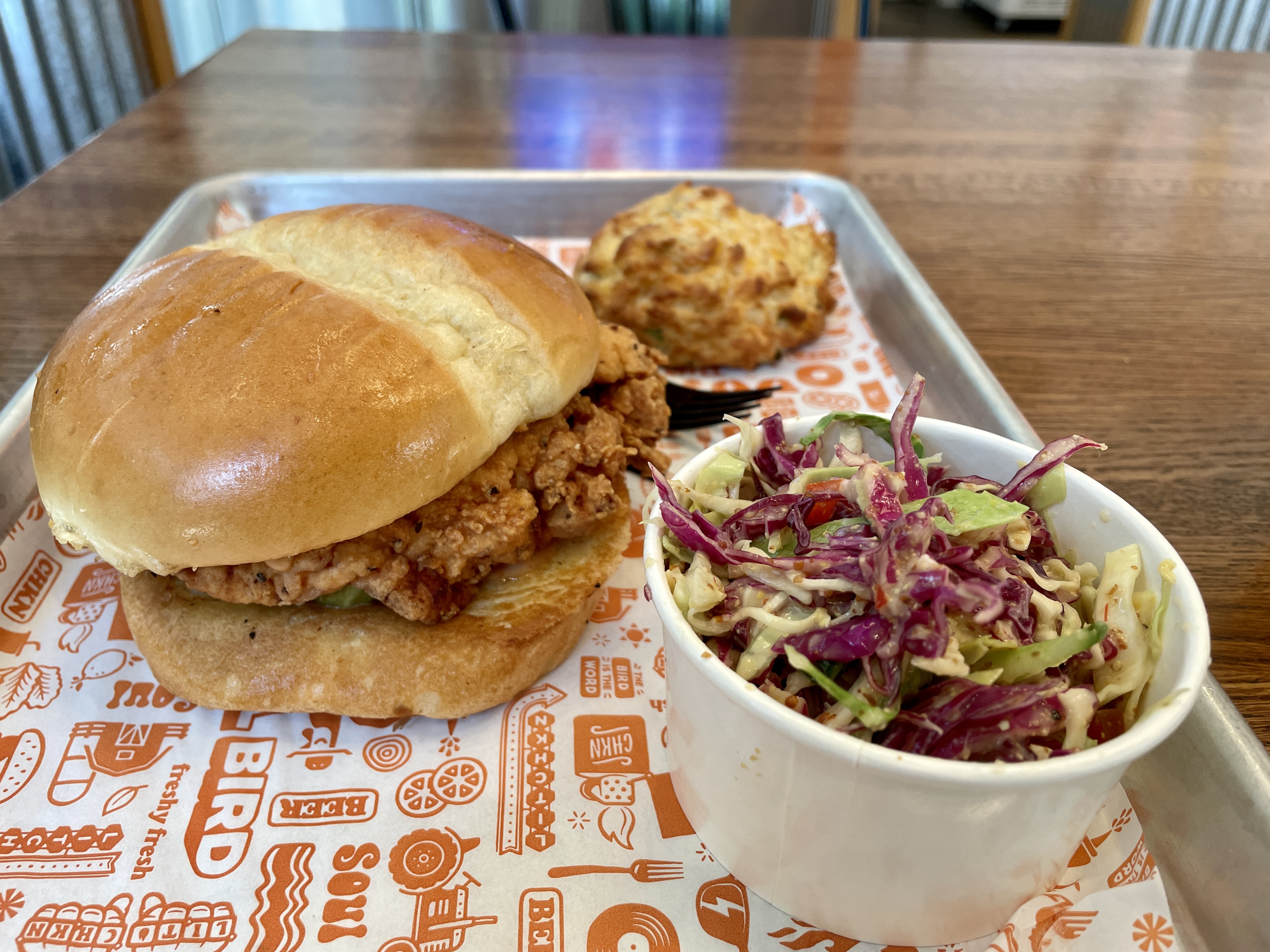 On a silver tray lined with white and orange paper sits a fried chicken sandwich on a brioche bun, a paper container of cole slaw with purple and green cabbage, and a biscuit.