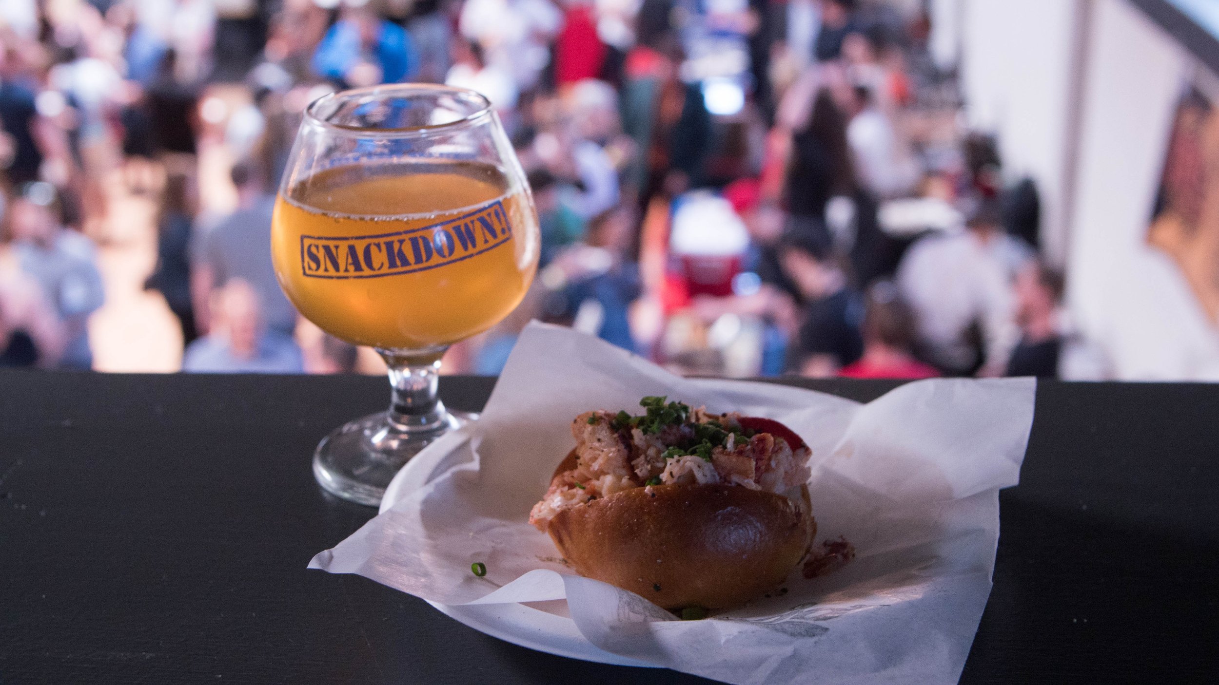 A Belgian beer glass sits next to a small lobster roll with a crowd in the background.