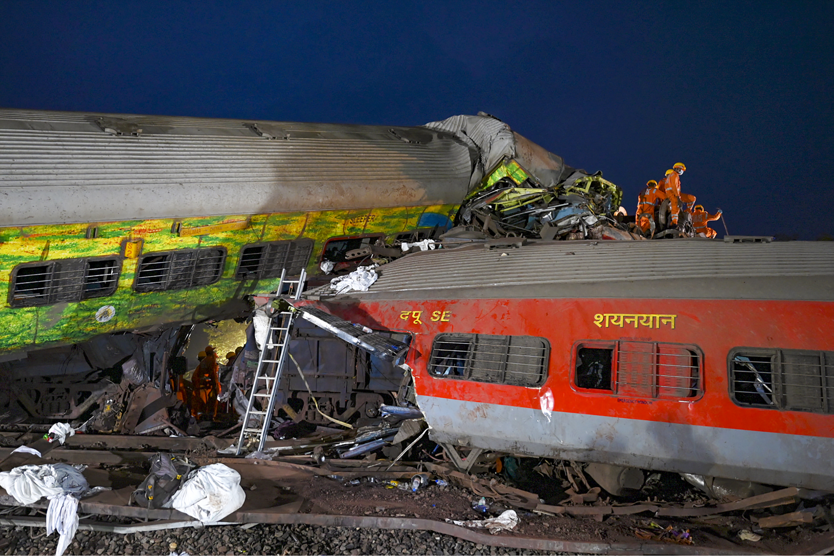 Two train carriages crushed together, one on top of the other, surrounded by debris. A ladder is propped up against the side of the train. Rescue workers in orange jumpsuits and yellow helmets stand on top of the train car. 