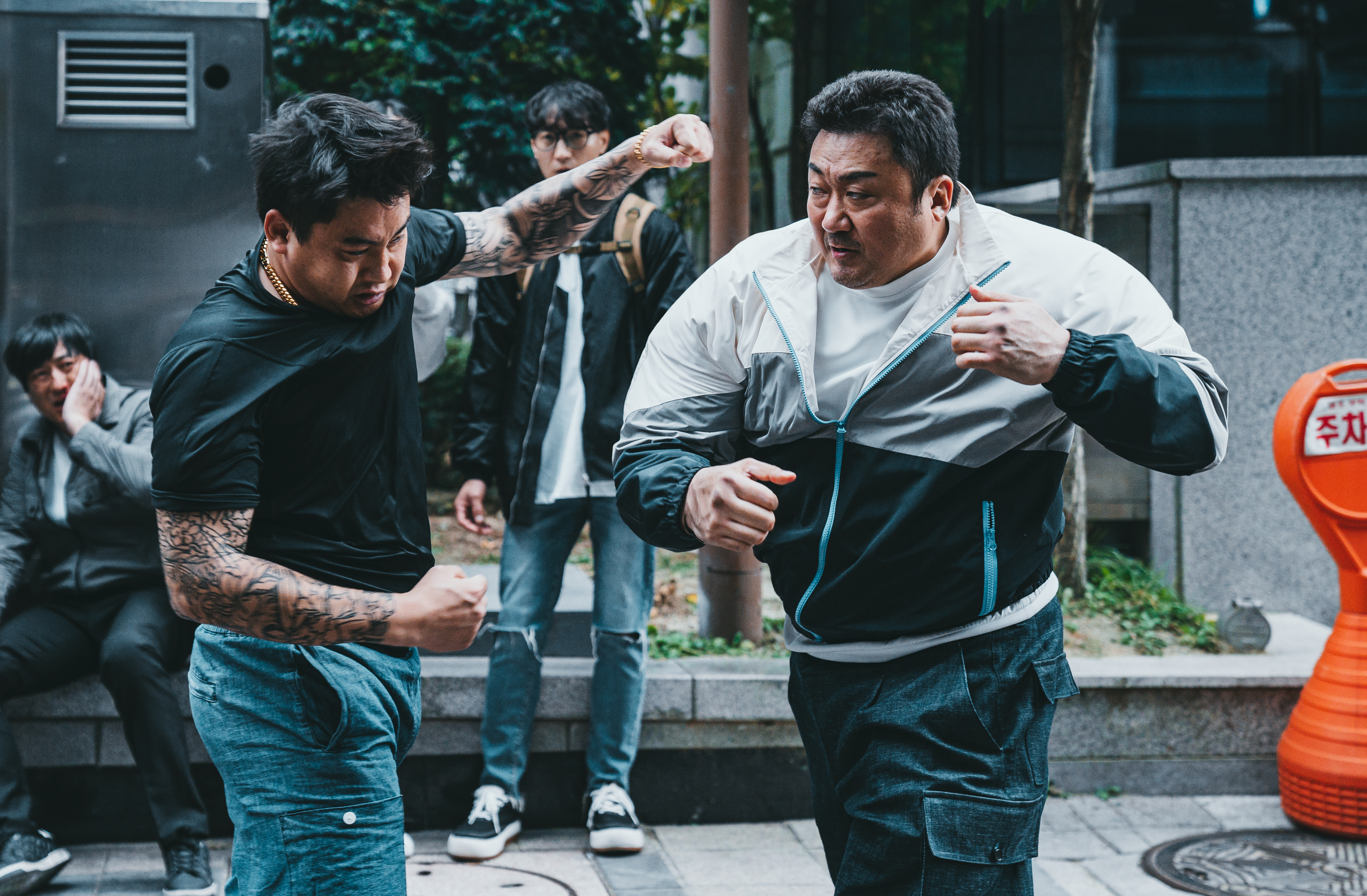 Ma Dong-seok, wearing a track suit, punches a young person in the stomach in The Roundup: No Way Out. The person being punched reacts as if they’ve been hit by a truck.