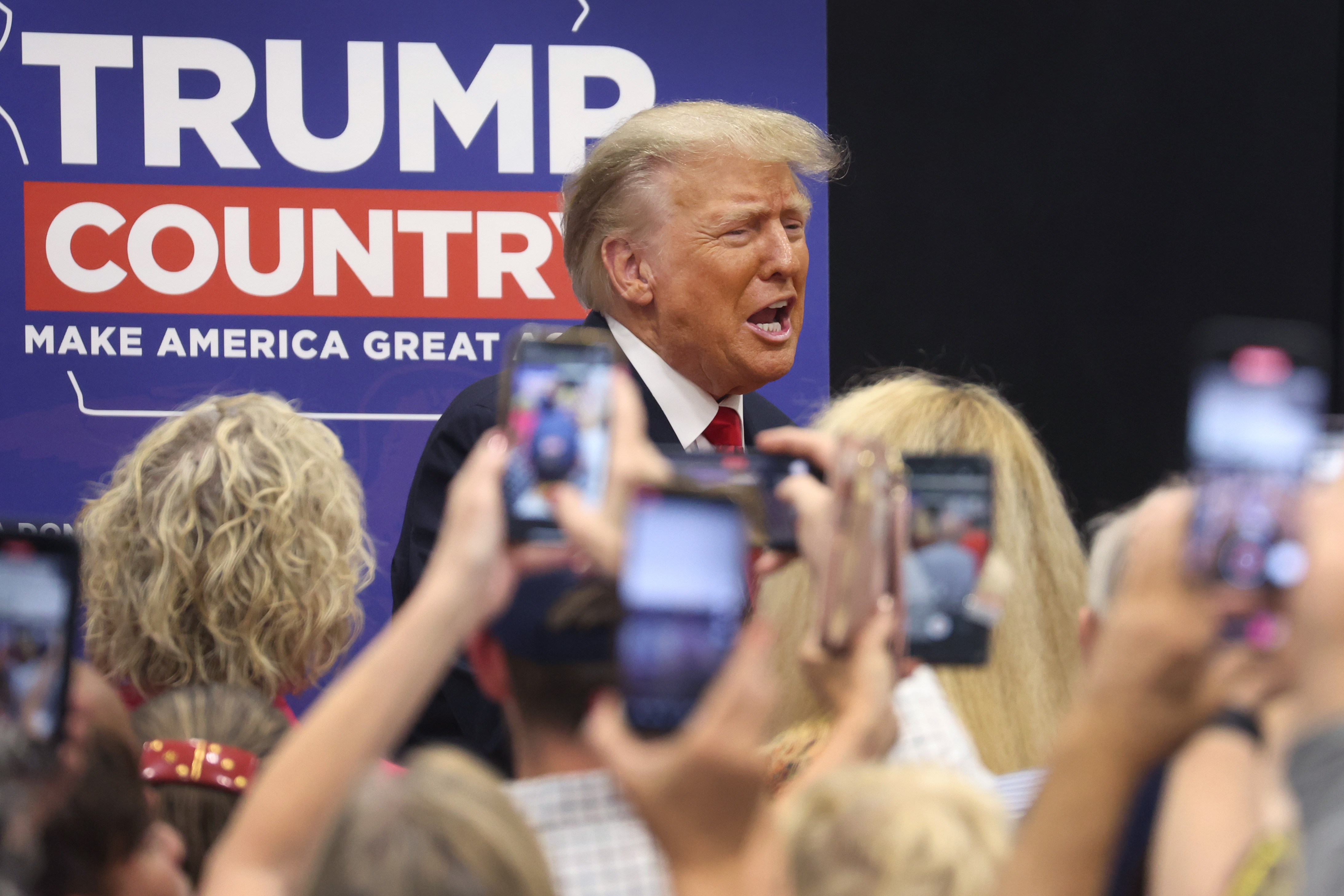 Donald Trump stands in front of campaign sign that says “Trump Country: Make America Great Again.” A crowd of onlookers take photos and videos with raised smartphones.