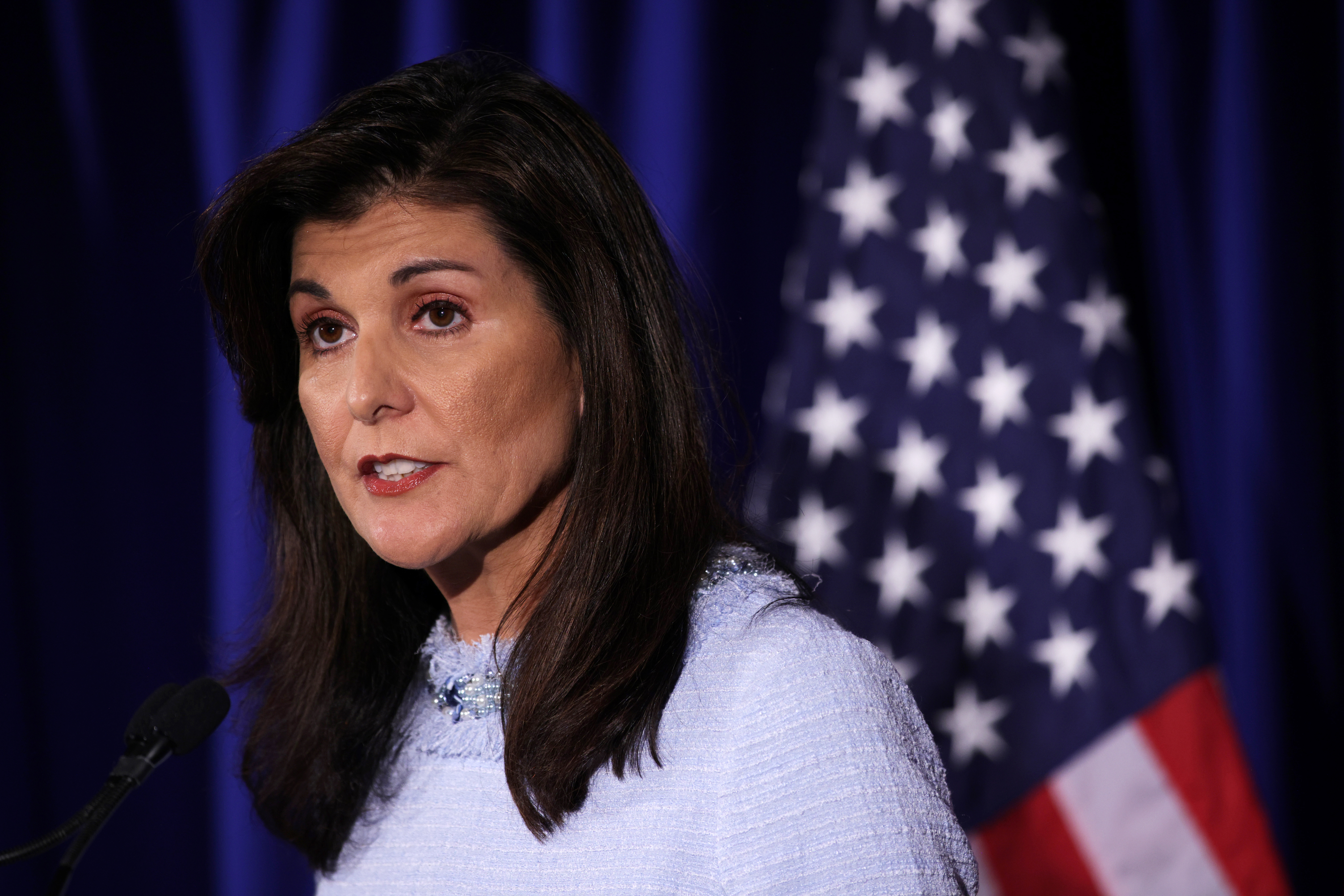 Nikki Haley, a woman in her 50s with long dark hair wearing a lavender dress, speaks with a serious expression. Behind her is a US flag against a blue backdrop.