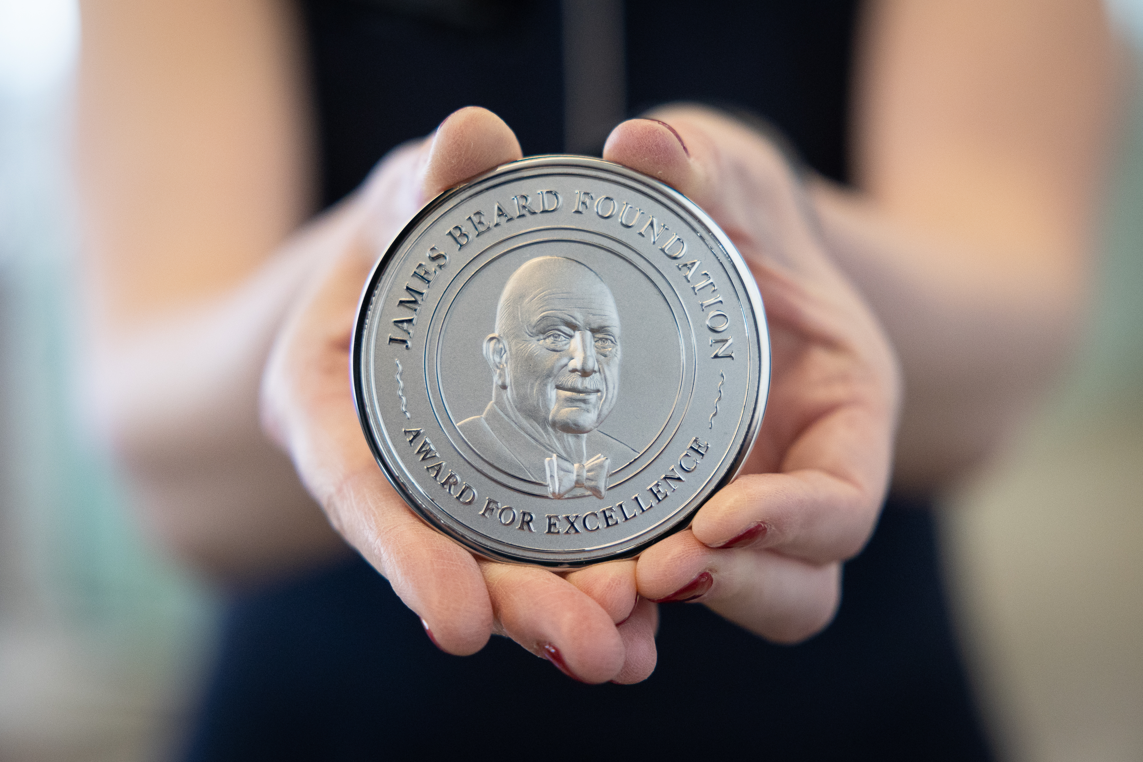 A woman holds a silver James Beard Foundation medal towards the camera, in the center of the shot.