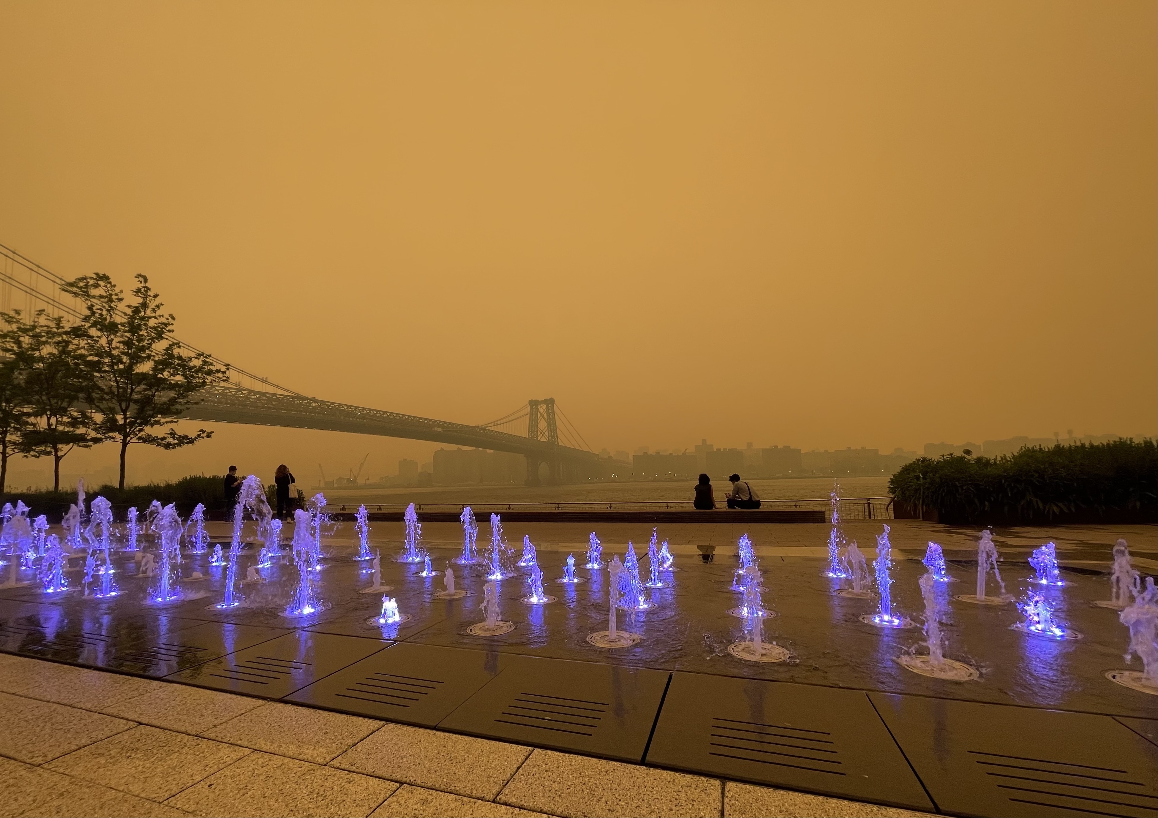 A smoky haze casts an orange glow on uplit  fountains in the foreground and a bridge in the distance.
