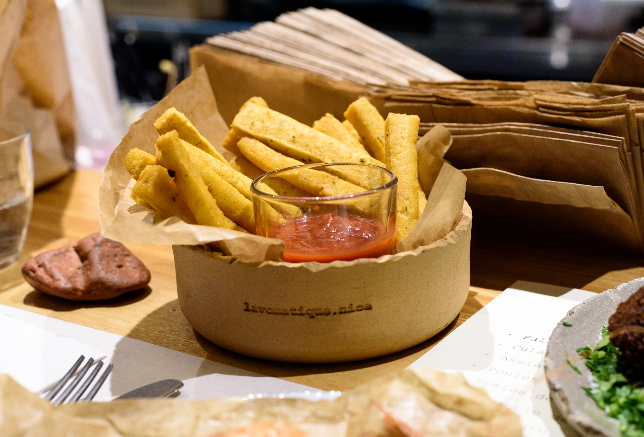 A clay bowl etched with the name Lavomatique, lined with paper and stuffed with sticks of fried panisse, along with a deep red dipping sauce. 
