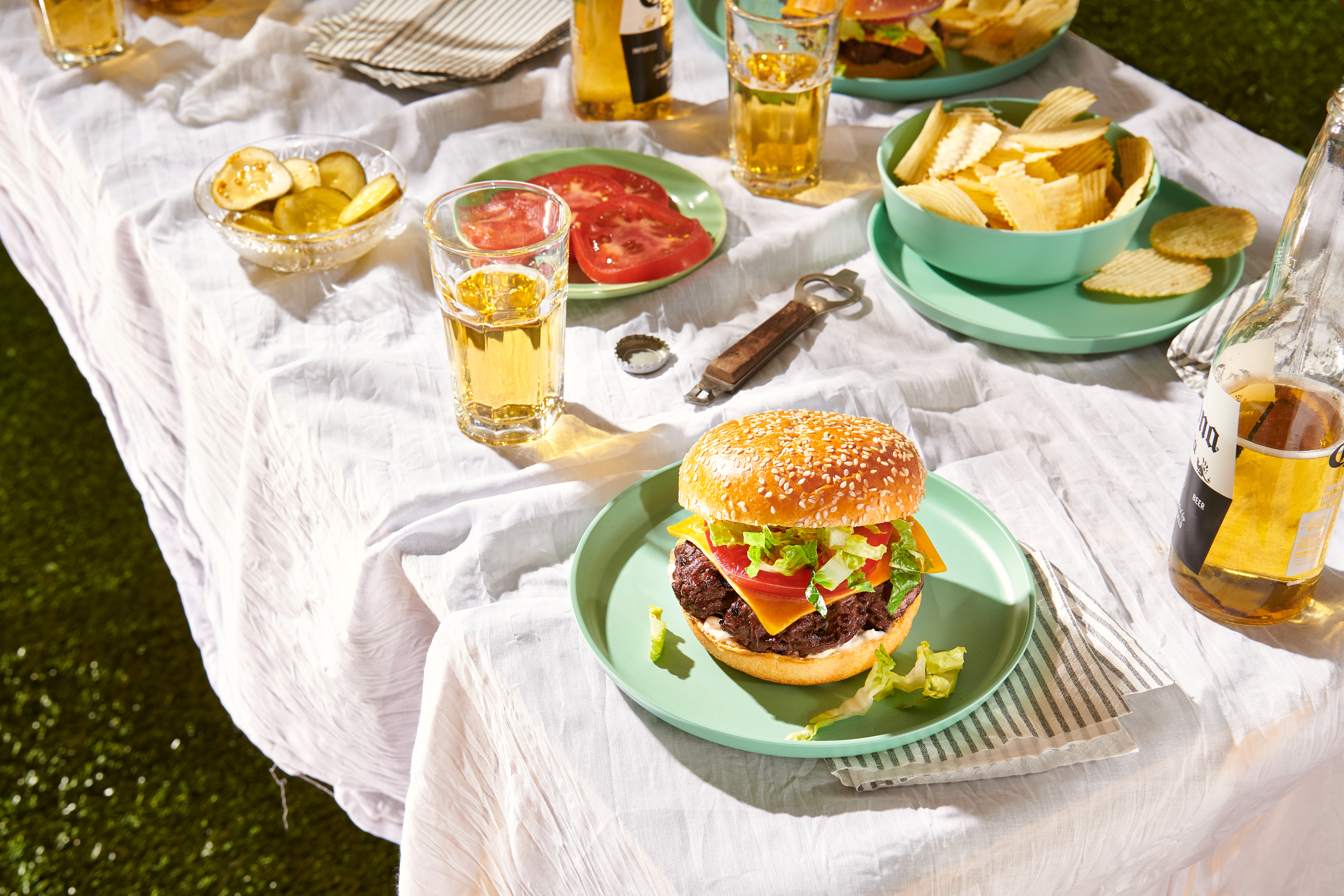 A burger topped with cheese, lettuce, and tomato sits on a table set with plates of tomato slices and chips and bottles of beer.