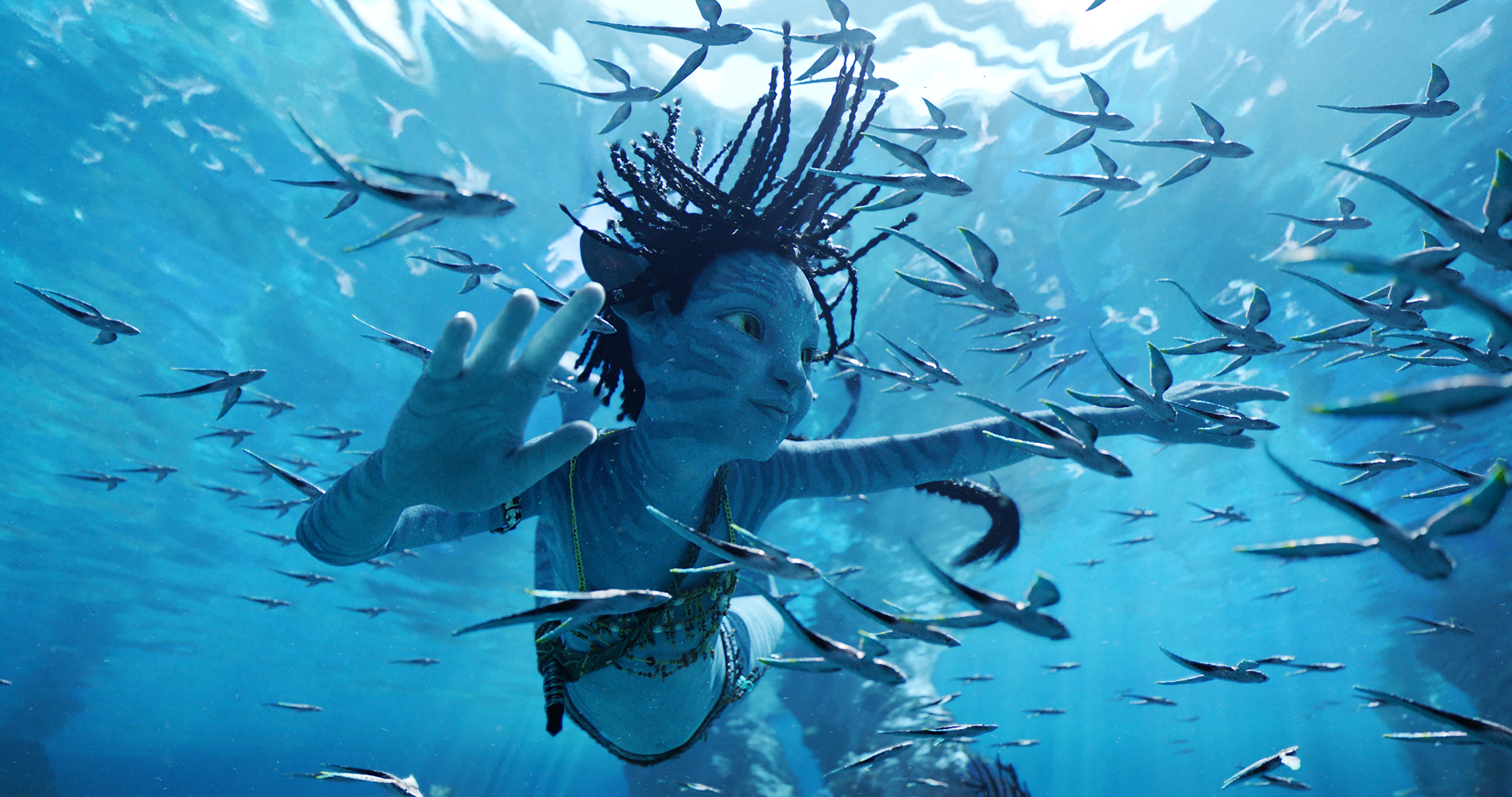 A young Na’vi child named Tuk (Trinity Bliss) swims underwater with her braids floating around her as she examines a school of tiny fish in Avatar: The Way of Water
