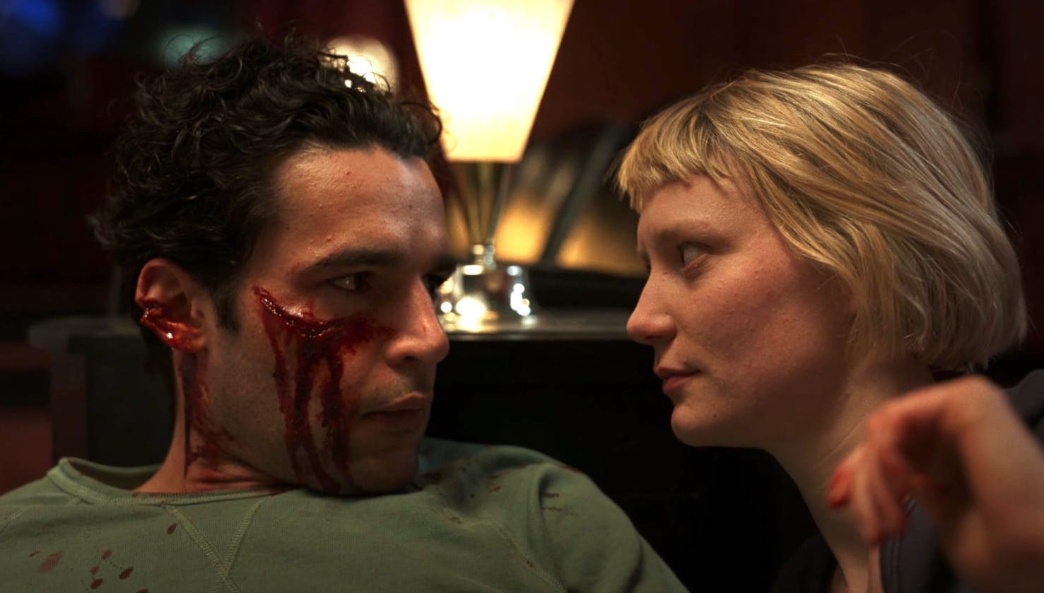 A man in a green shirt bleeds out his eye while sitting on the couch looking at a young blonde woman in Piercing