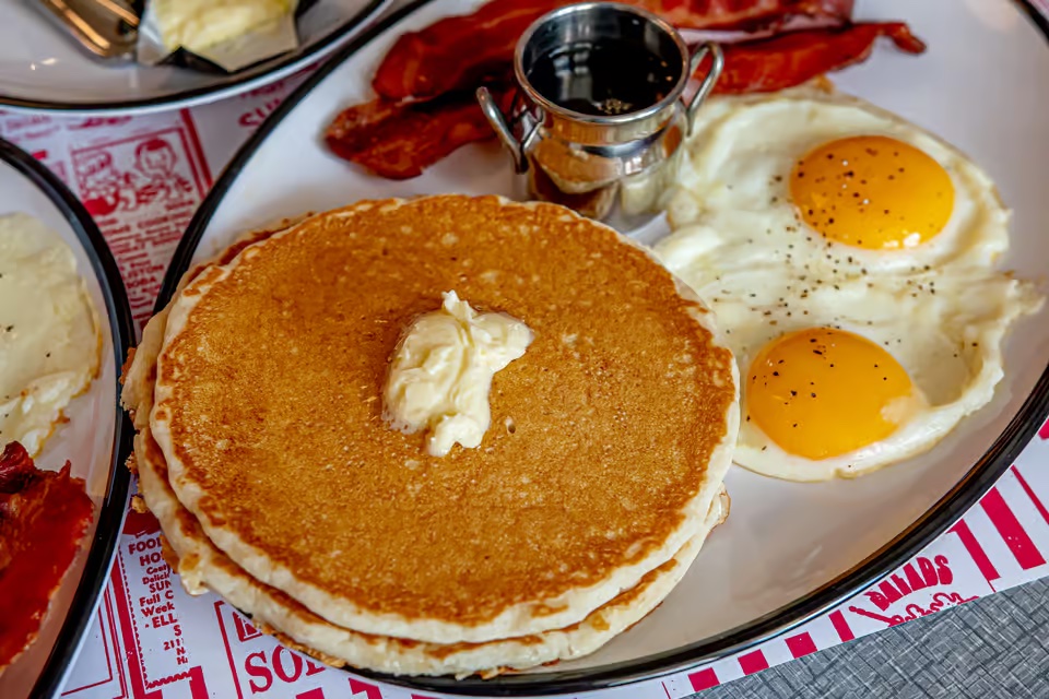 A closeup of a classic diner breakfast plate including two pancakes with butter on top, two sunny side up eggs, a metal container of syrup, and two slices of bacon.