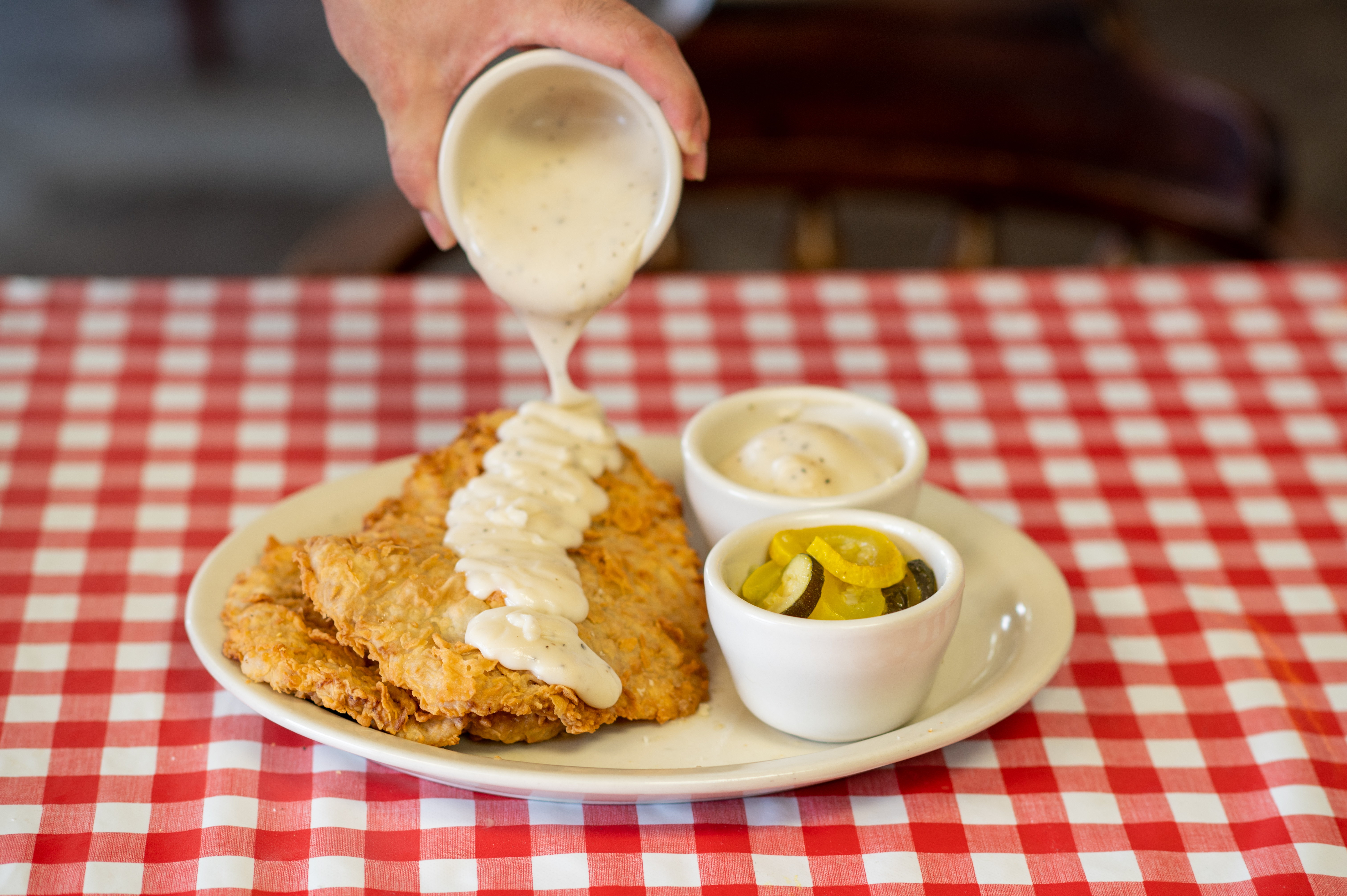 A table with a red checkered tablecloth holds a plate of chicken-fried steak with cream gravy, with two ramekins of mashed potatoes and squash. Above it, a hand holds a third ramekin with gravy that is still pouring out.