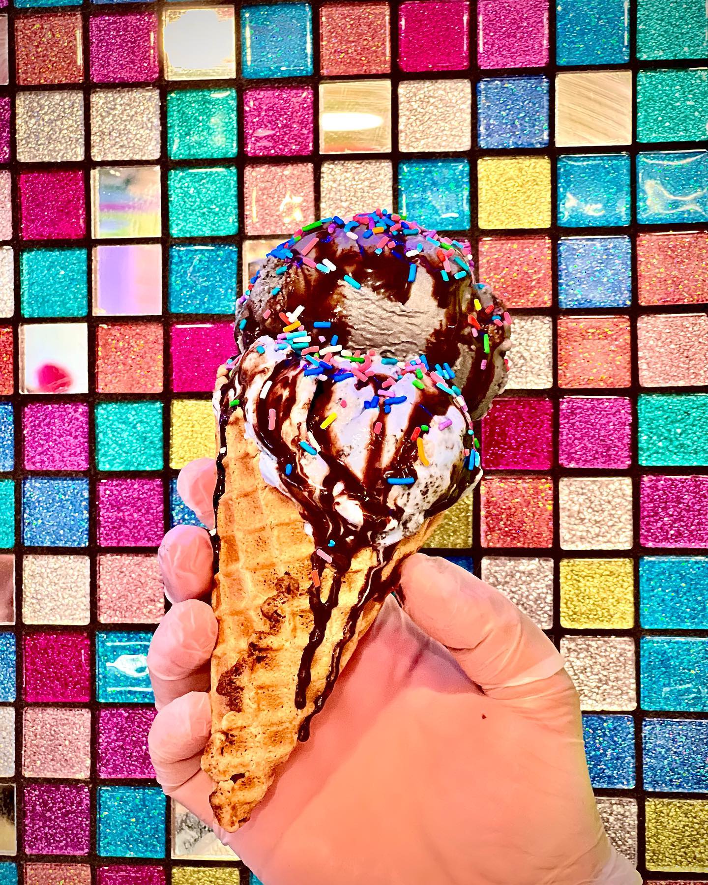 Someone holding up a waffle ice cream cone with chocolate sauce and sprinkles in front of a colorful tiled background.