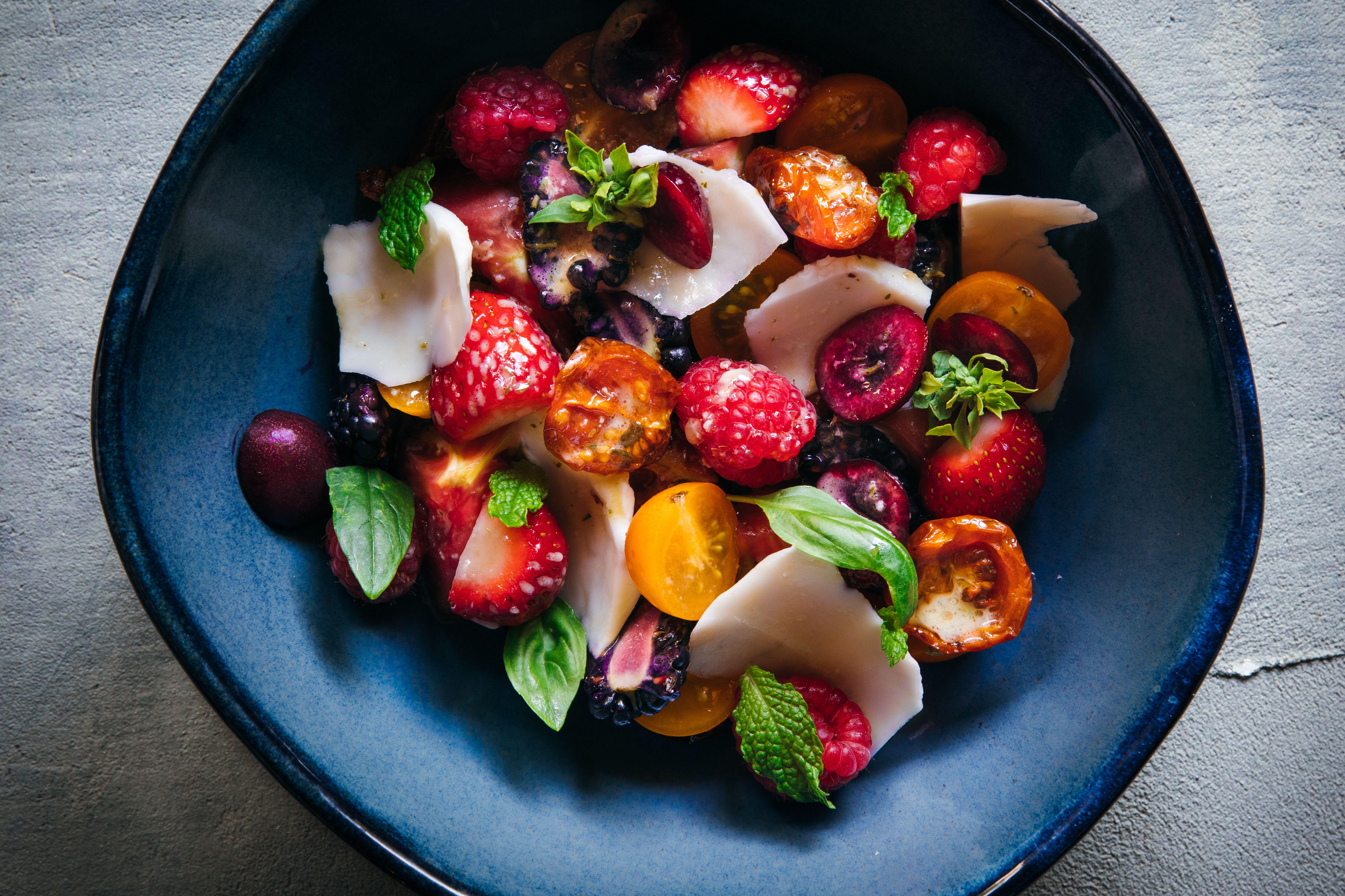A blue bowl is filled with tomatoes, strawberries, raspberries, and cherries, with chunks of young coconut and fresh herbs. This is one of the summer salads at Kann.