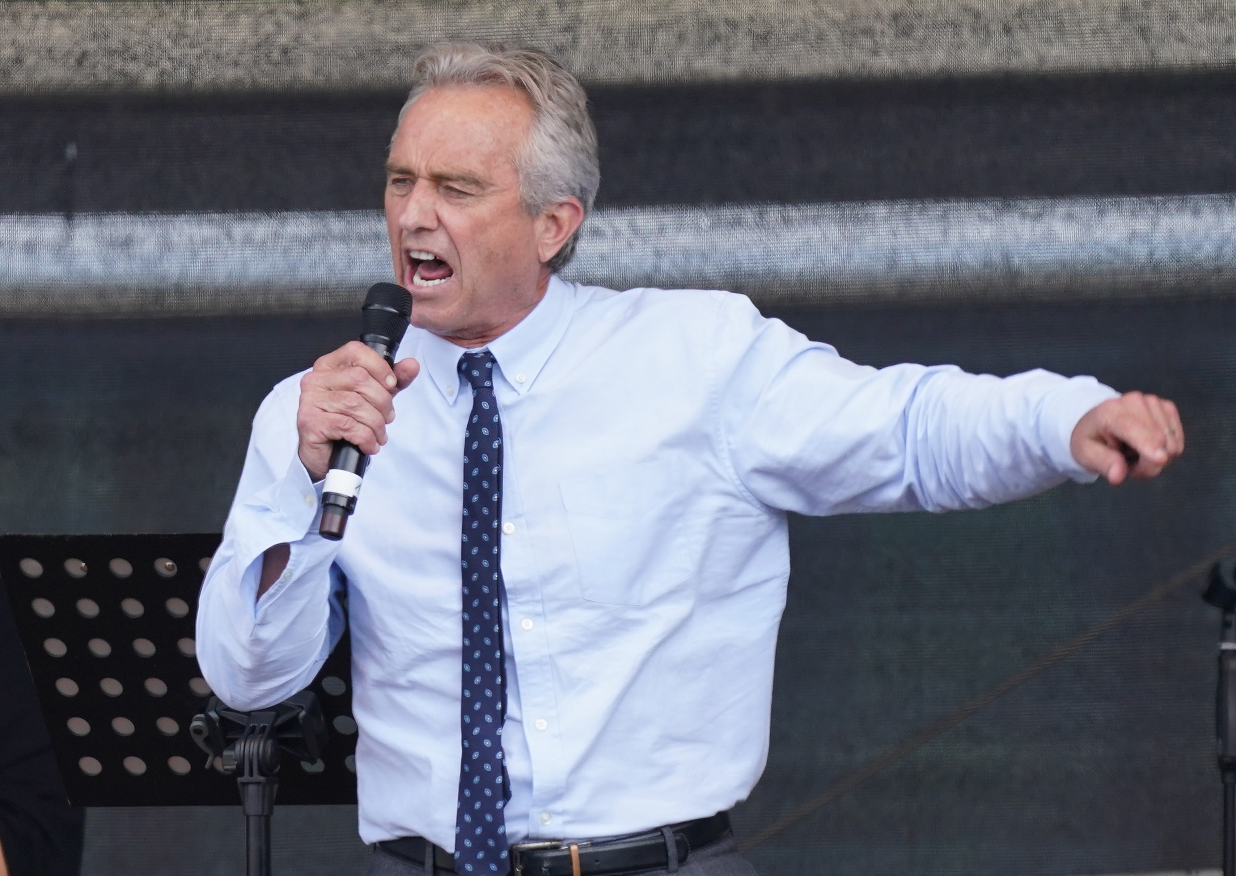 Kennedy, in a white shirt and slim blue tie, speaks emphatically into a handheld microphone, his other arm extended in a gesture.