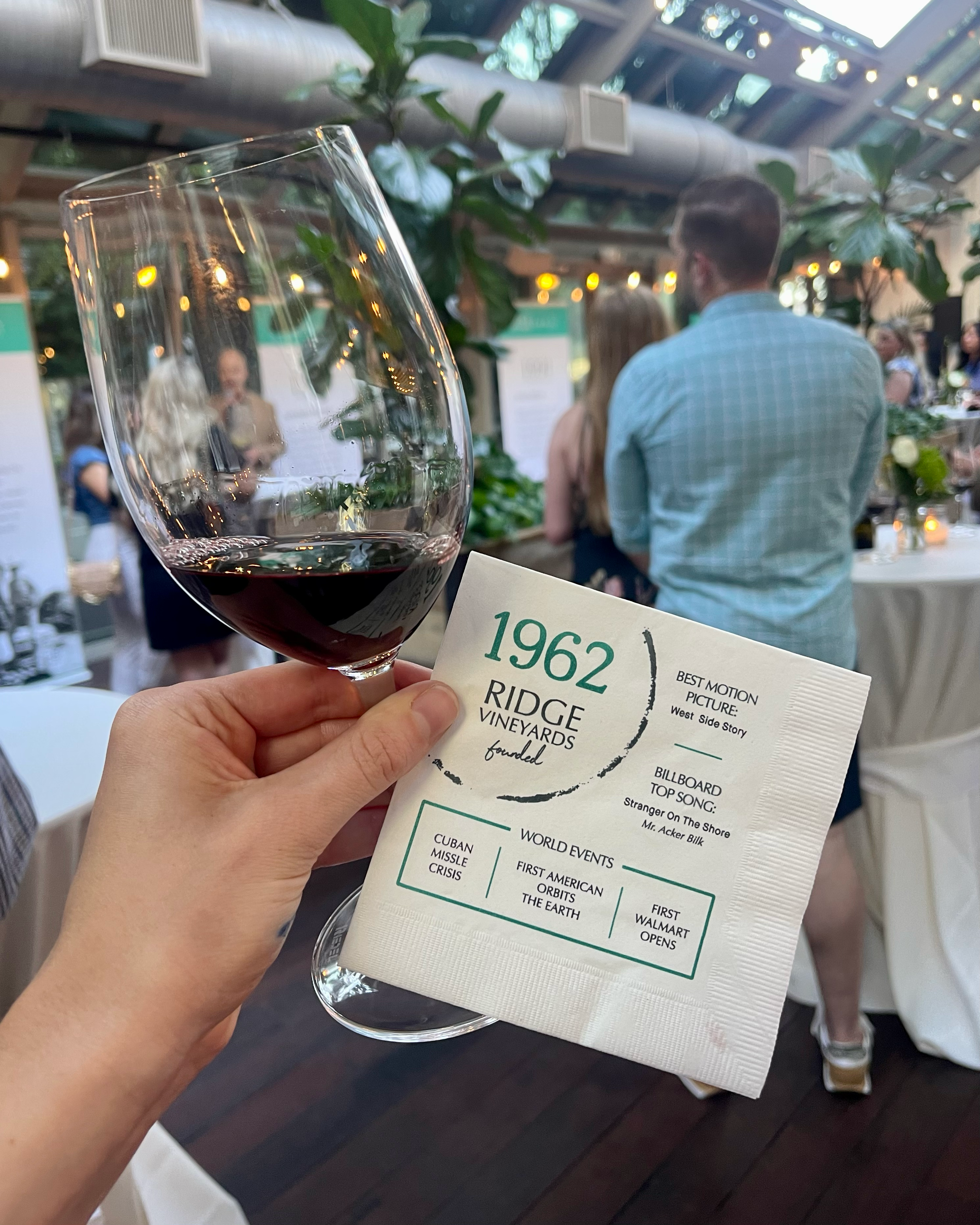 In a busy room, a hand holds a glass of red wine in front of the camera. It also holds a napkin embossed with “1962 Ridge Vineyards,” and a list of notable events from that year.