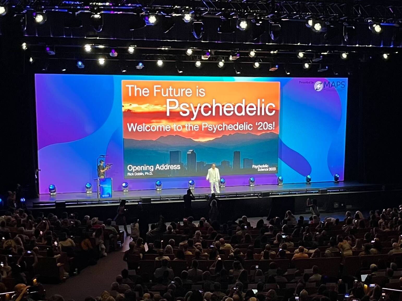 A person onstage in front of a large colorful screen that reads “The future is psychedelic.”