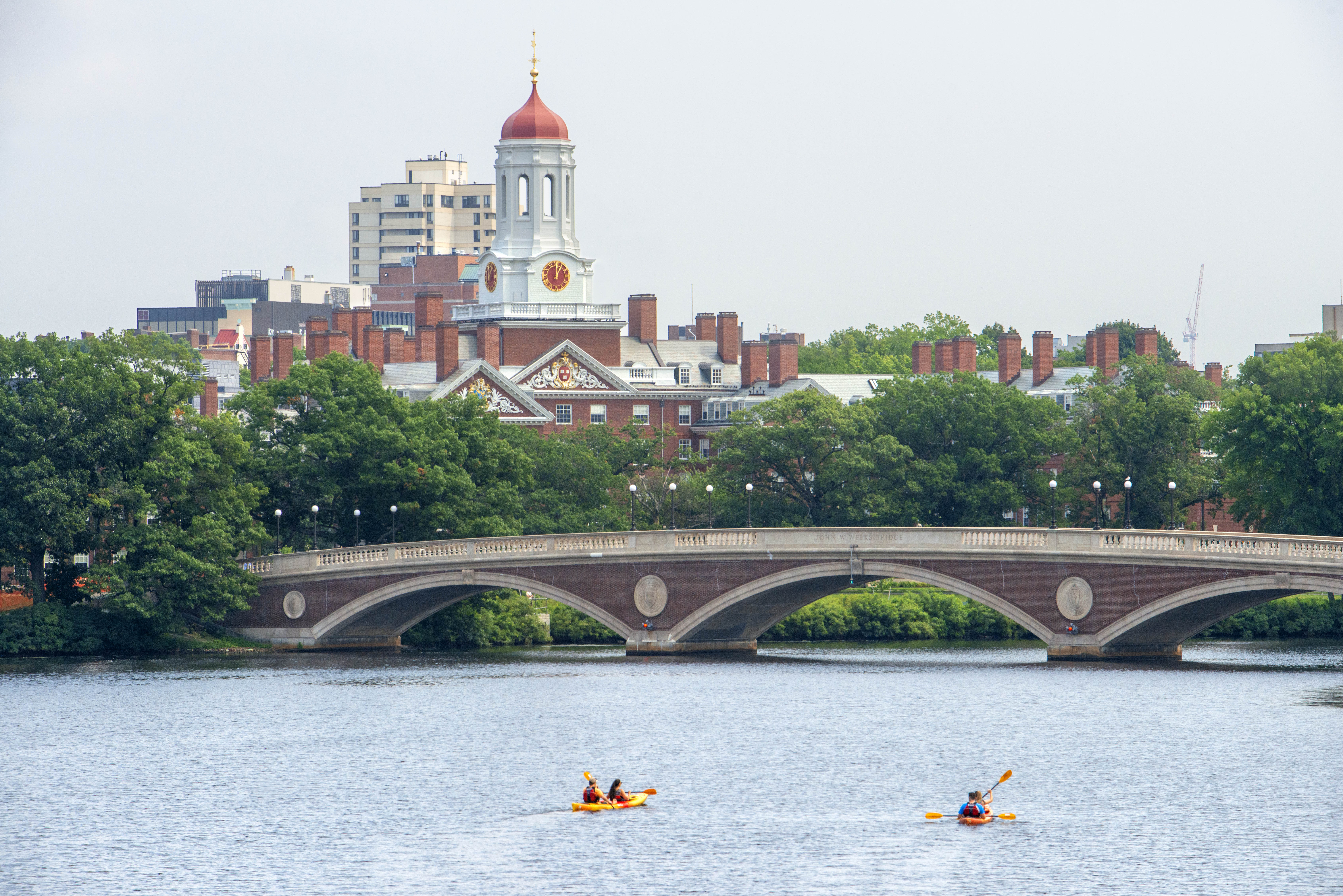 A photo of a river in the foreground, with kayakers and a bridge. In the backround is a clock tower and buildings of Harvard University campus.