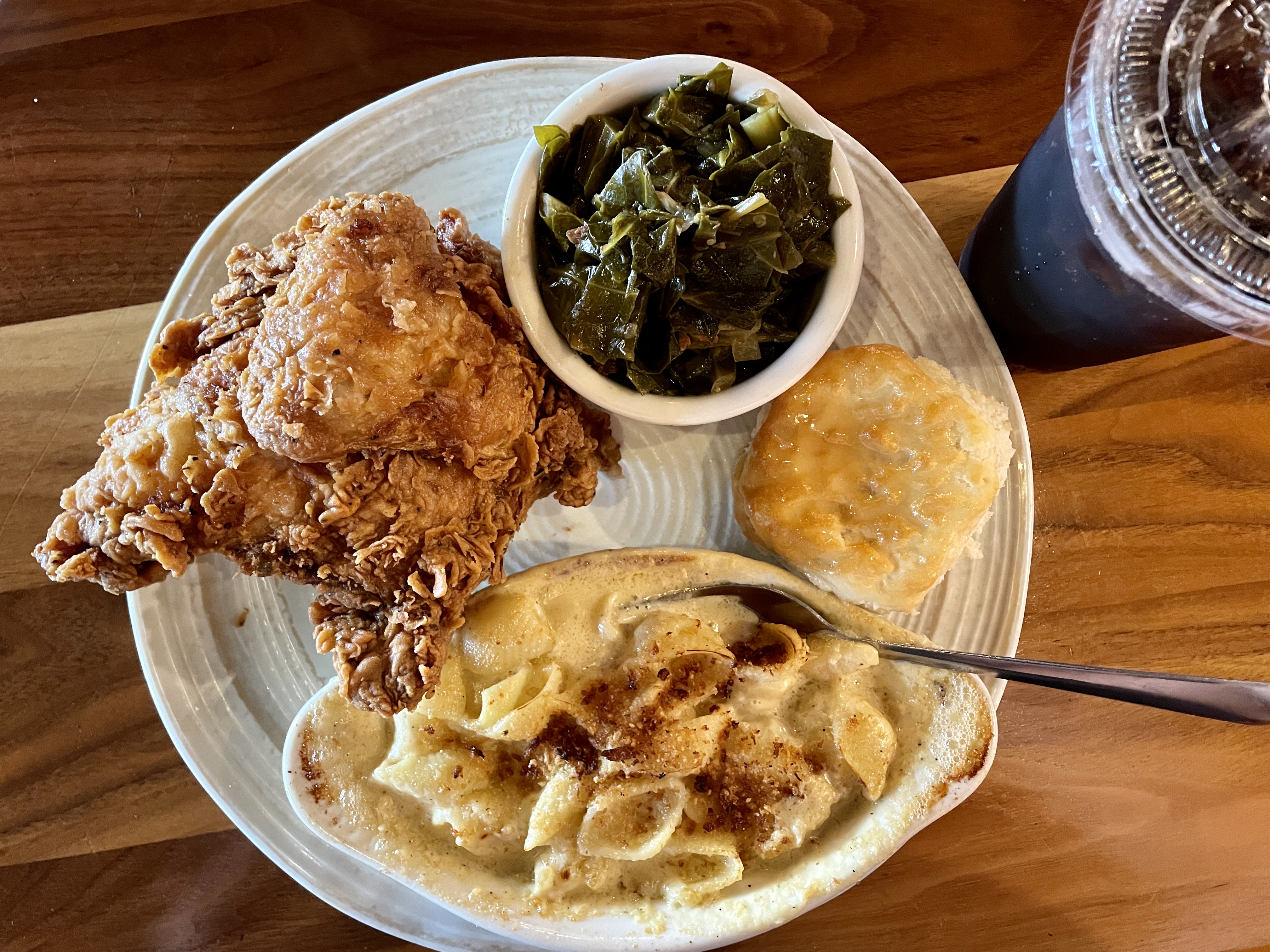 A thick platter holds two pieces of fried chicken, a small bowl of greens, and a dish of mac and cheese with a fork in it. To the right is a plastic glass of soda.