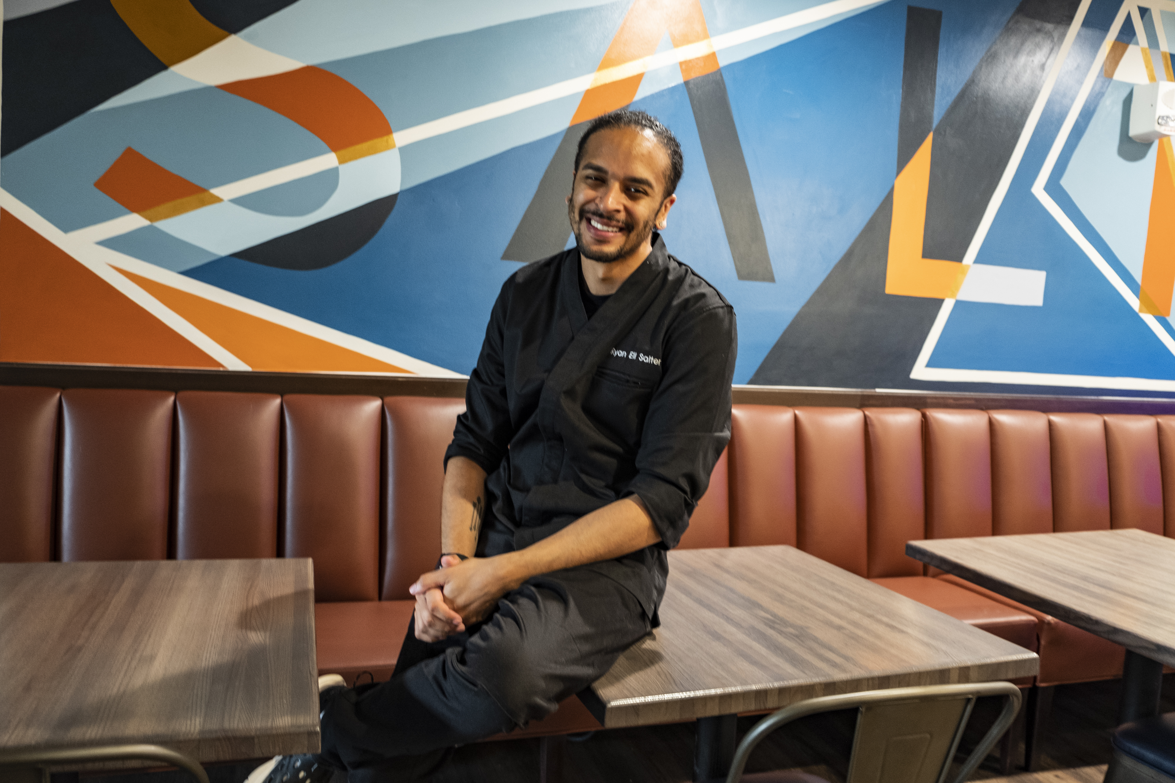 A man with dark hair pulled back, wearing a black chef jacket and black pants leaning on a table in front of a mural at Salt &amp; Ko. in Southfield, Michigan.