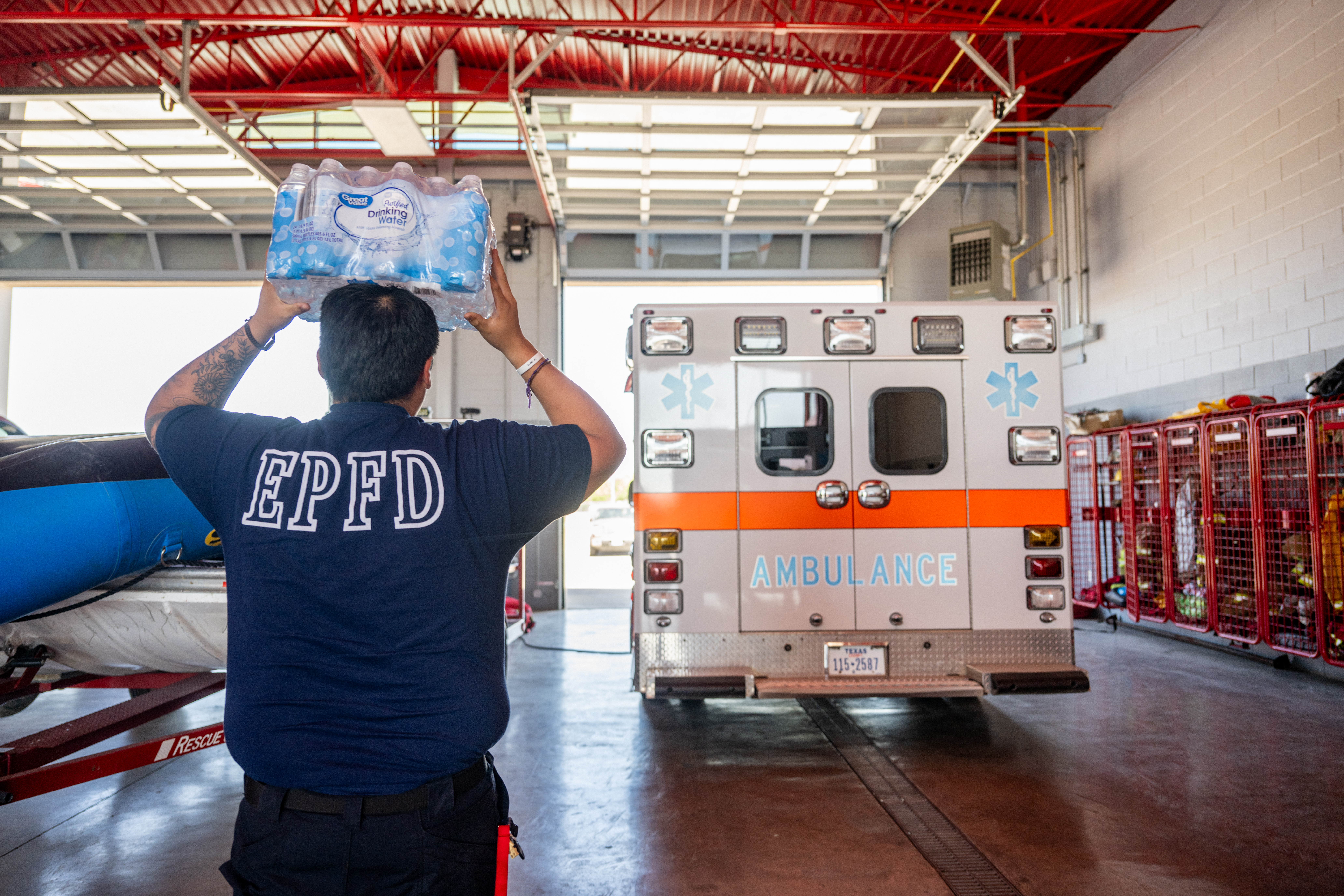 An EMT with his back to the camera holds a case of water above his head. An ambulance is seen in the background. 