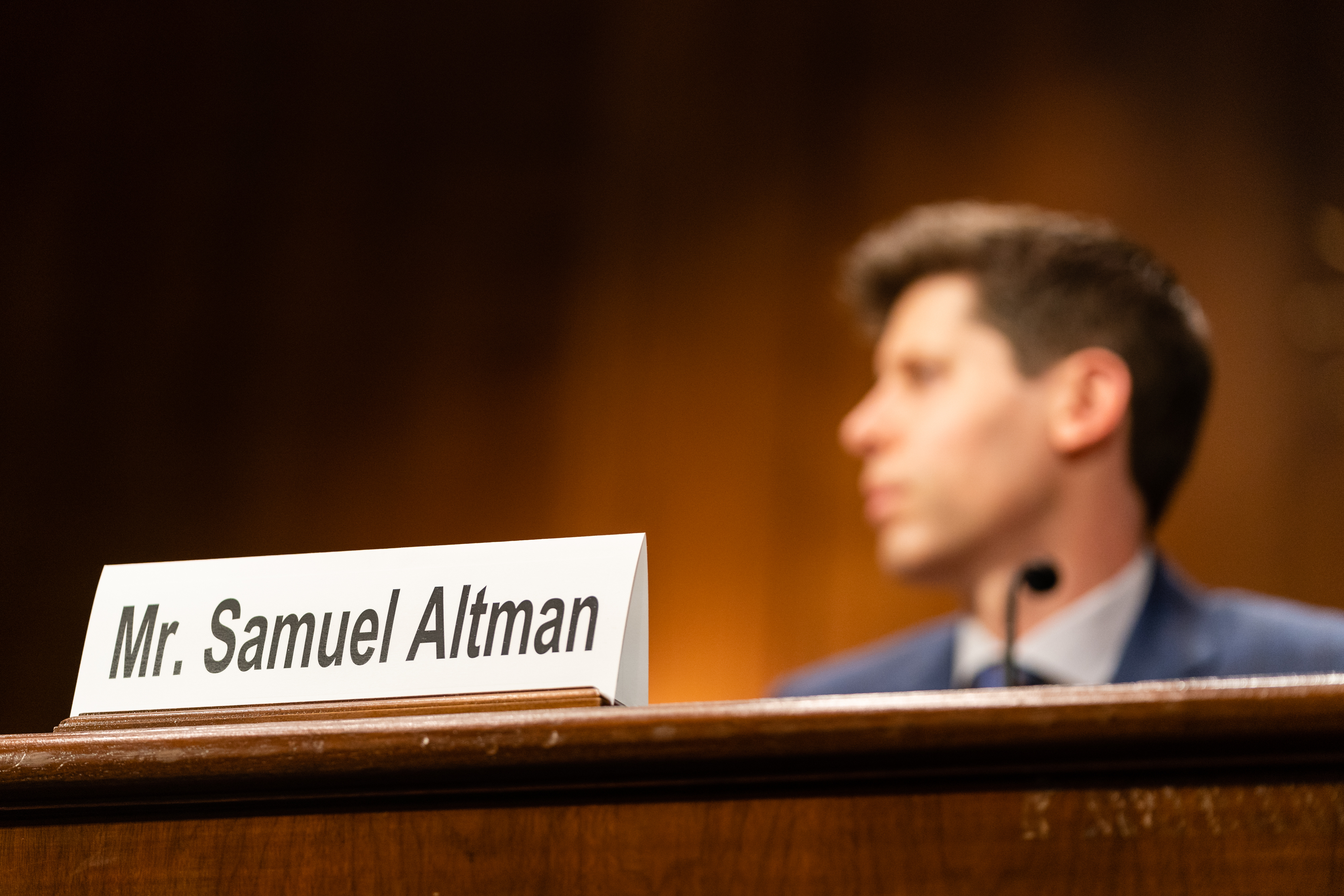 A nameplate reading Mr. Samuel Altman in the foreground. Altman is seen in the background. 