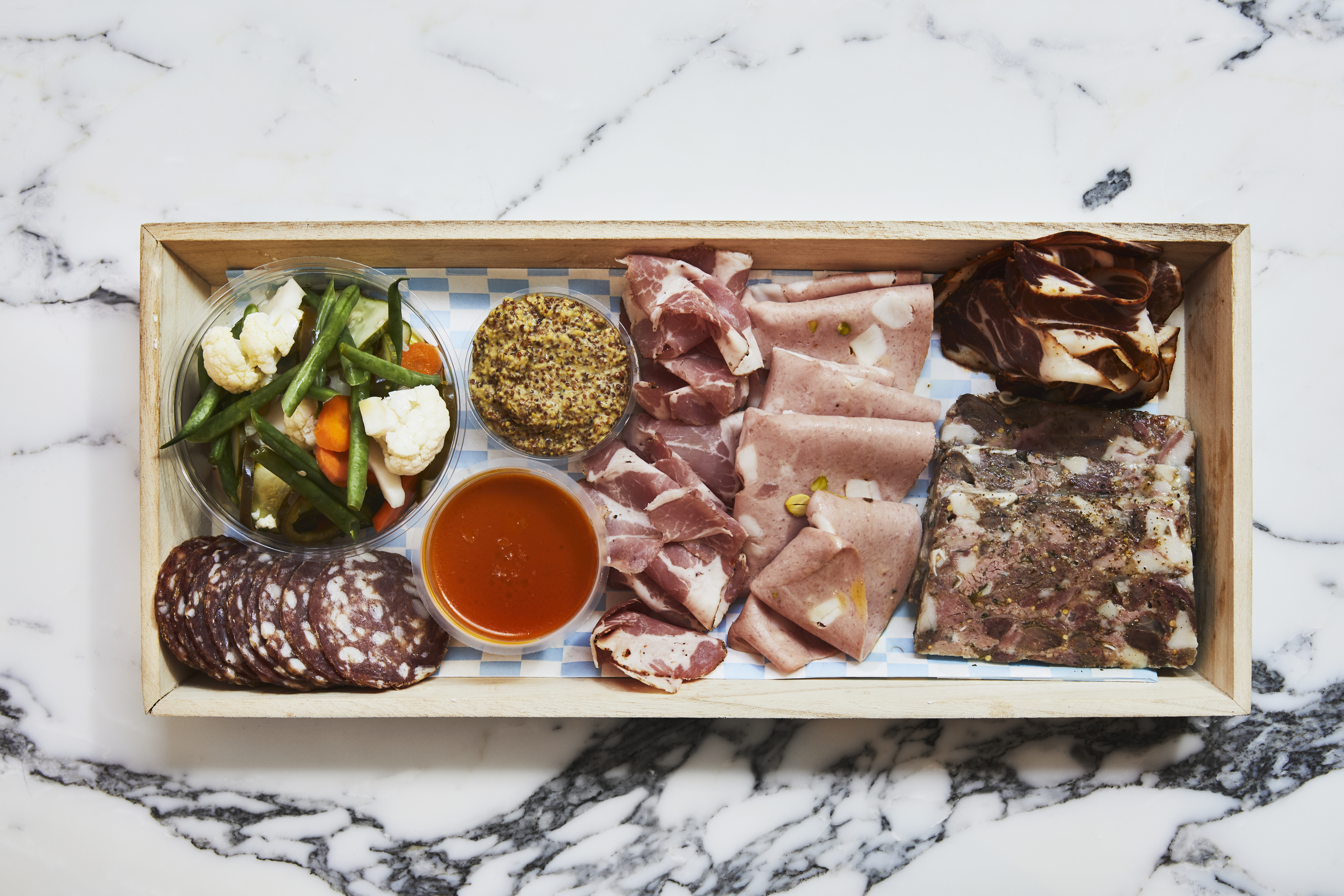 A wooden box on a marble counter filled with pickled veggies and cured meats