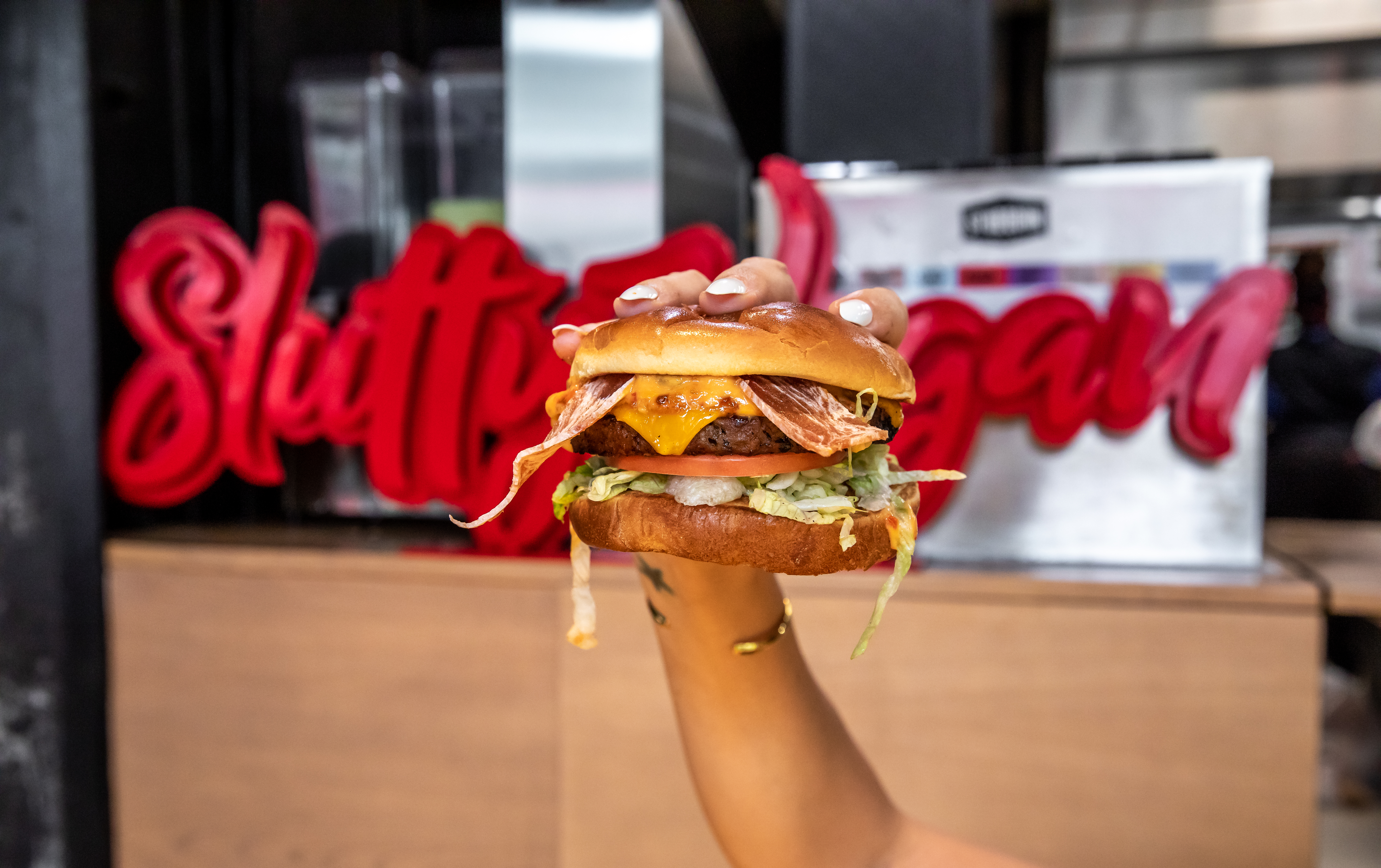 A woman’s hand holds up a vegan burger loaded with toppings, with the words “Slutty Vegan” behind it in red script.