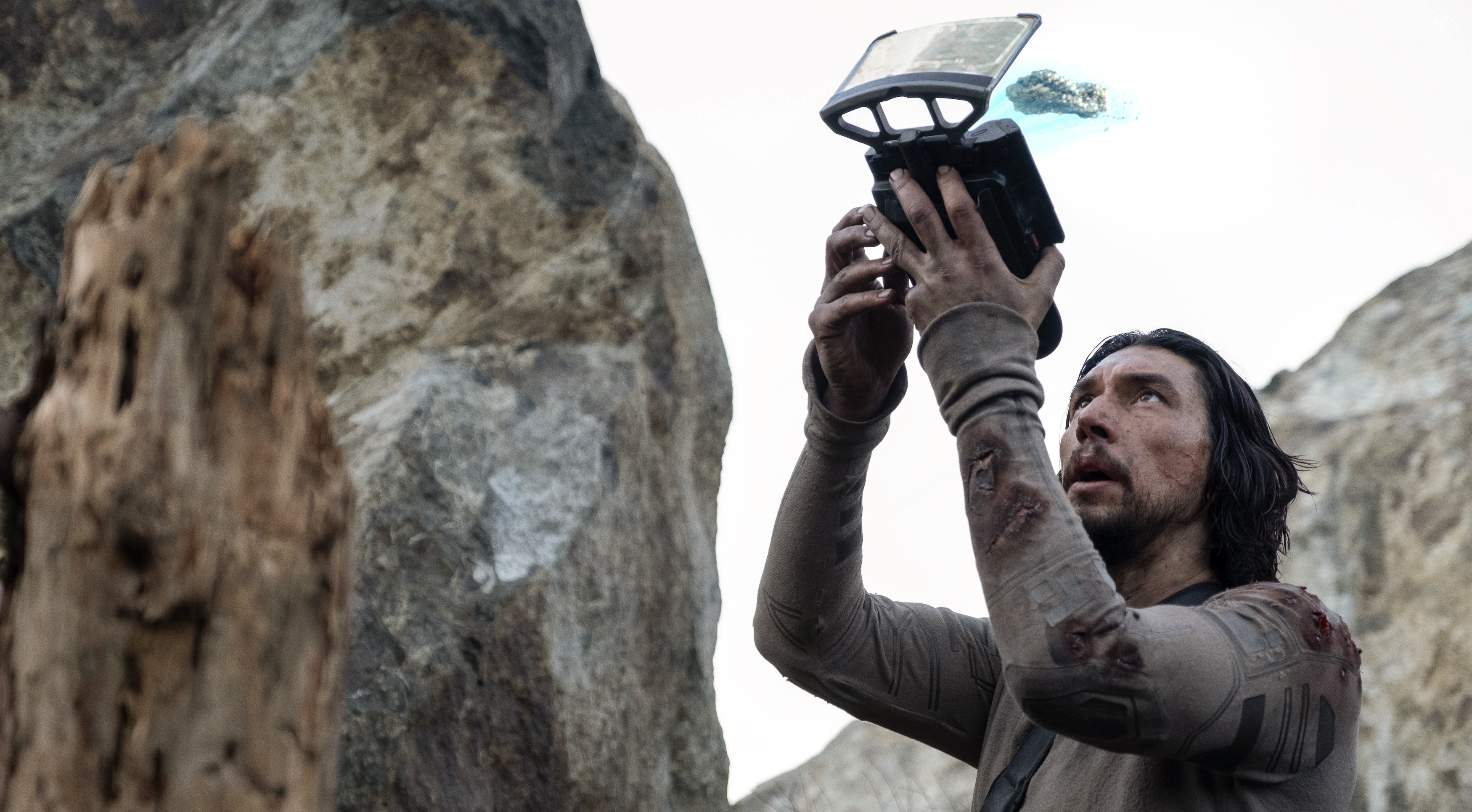 Mills (Adam Driver), a space trucker stranded on Earth, looks grubby and wounded as he stands by a giant rock and holds up a small, boxy device thats projecting a small hologram of a ship image in 65